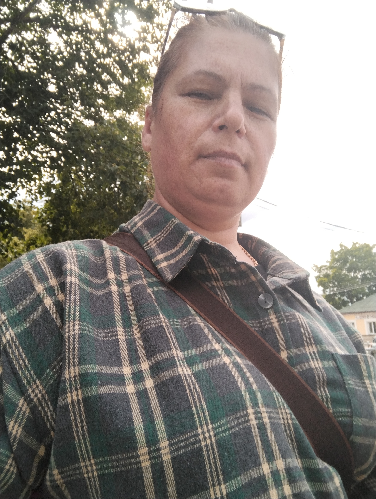A woman with light skin, freckles, and pulled-back light brown hair is posing for a selfie. She is wearing a green, navy, and tan plaid shirt over a gold chain necklace. A brown strap from a bag crosses her chest from her right shoulder to her left hip. She is outdoors, with trees and a bright sky visible in the background. In the far background, a building with windows and a slanted roof can be seen. The image appears to have been taken on a street in Kingisepp, Russia, given the architecture of the building.