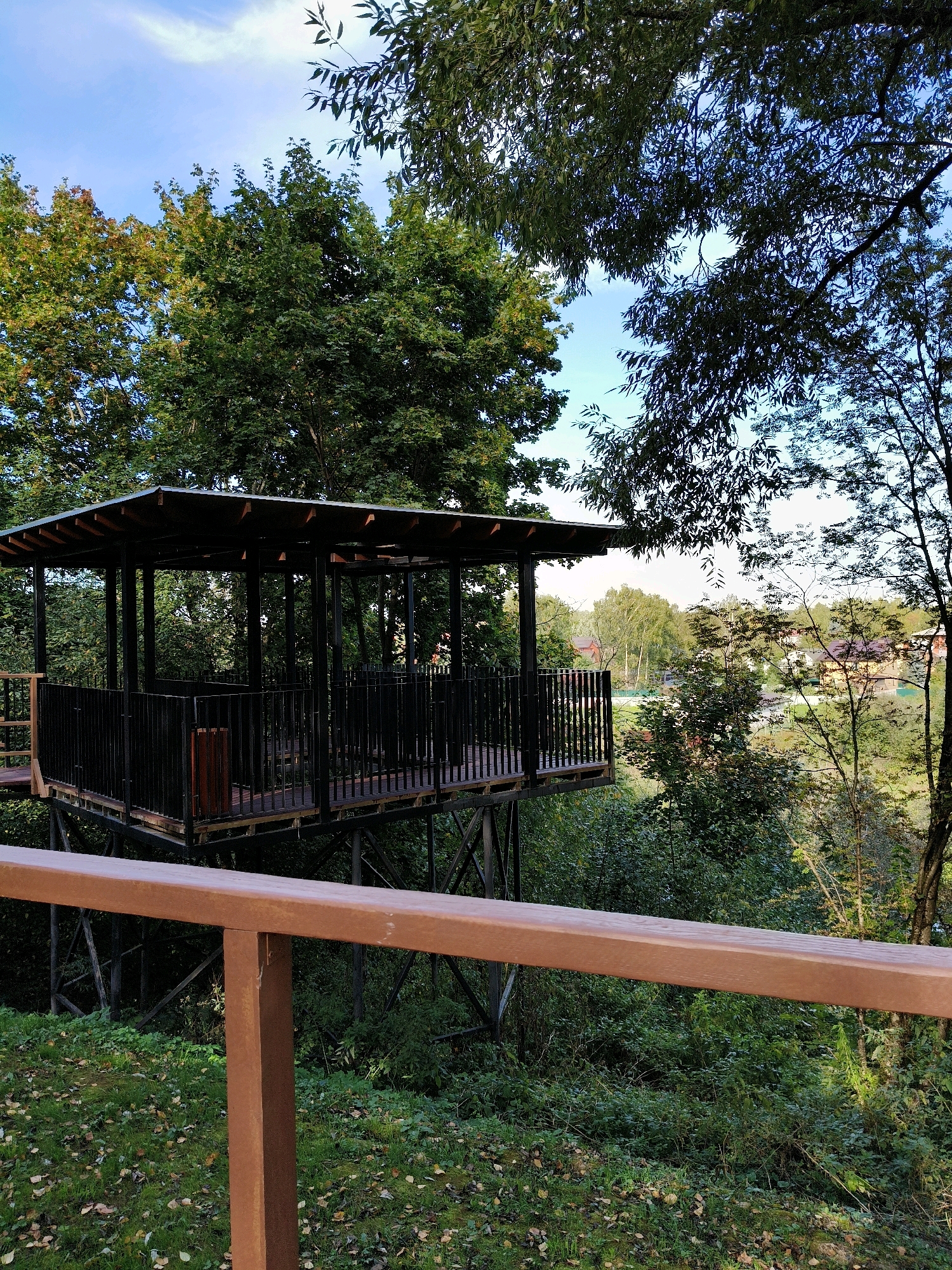 The image depicts an elevated, covered wooden pavilion with black metal railings, situated within a lush, green, wooded environment. The pavilion is supported by a dark metal structure. In the foreground, a wooden railing, colored a reddish-brown, runs horizontally across the lower portion of the frame. Behind the pavilion, a residential area with houses and trees is visible under a blue sky with scattered clouds. The scene is set in the city of Naro-Fominsk, Russia, and appears to be a public recreational area, possibly a park or a viewpoint. No people are present in the image.