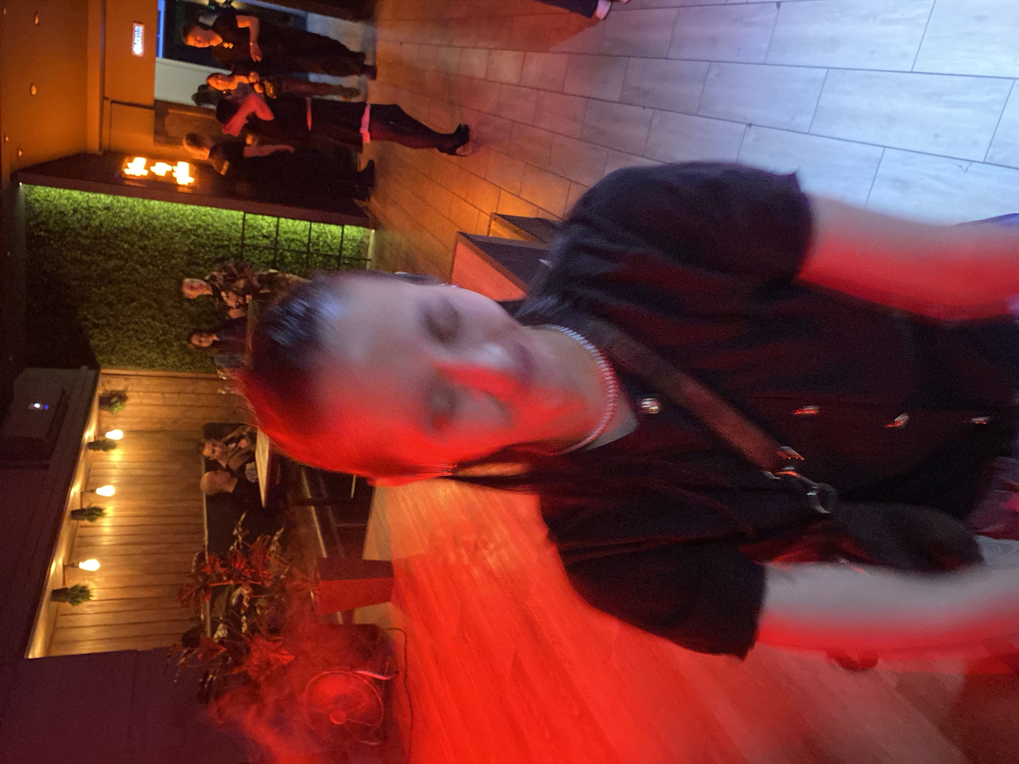 The media file, rotated 90 degrees clockwise, captures an indoor or semi-outdoor social venue, likely a restaurant or bar, at night in Votkinsk, Russia.
In the foreground, a woman's face is prominent, angled towards the left. Her eyes are closed or looking down, and her features are bathed in a strong red light emanating from below. She wears a dark top and a silver necklace, with a strap visible over her shoulder. A small, dark fan rests on the red-lit wooden floor near her.
Further into the background, a wall covered in artificial green foliage stretches across the middle of the scene. Above this foliage, a digital display shows illuminated orange characters, resembling "4-4". To the left of the foliage wall, a wooden surface is adorned with warm string lights featuring visible bulbs, and a few indistinct figures can be seen seated at tables. On the right side of the background, a light-colored tiled floor is visible, illuminated by a cooler blue light, with several individuals standing or walking, some near a set of stairs.
The overall atmosphere is dim and moody, characterized by a dynamic interplay of red, orange, and blue artificial lights, typical of an evening social gathering.