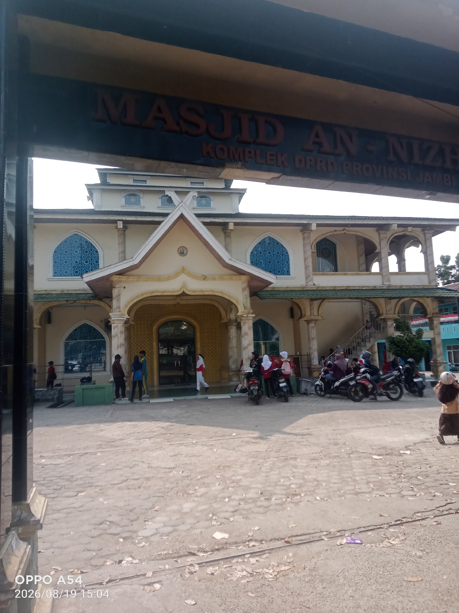 The image shows the exterior of Masjid An-Nizah, located within the Kompleks DPRD Provinsi Jambi in Jambi City, Indonesia. The mosque is a light-colored building with a prominent entrance featuring ornate yellow detailing and an arched doorway. Above the entrance, a large sign reads "MASJID AN-NIZAH" and below that, "KOMPLEK DPRD PROVINSI JAMBI". Several people are visible in front of the mosque, some standing and others on or near motorcycles parked on the paved area. The environment is outdoors, with a bright sky suggesting daytime. The ground appears to be paved with interlocking bricks or stones, with some debris and fallen leaves present. The overall scene depicts a public space in front of a mosque.