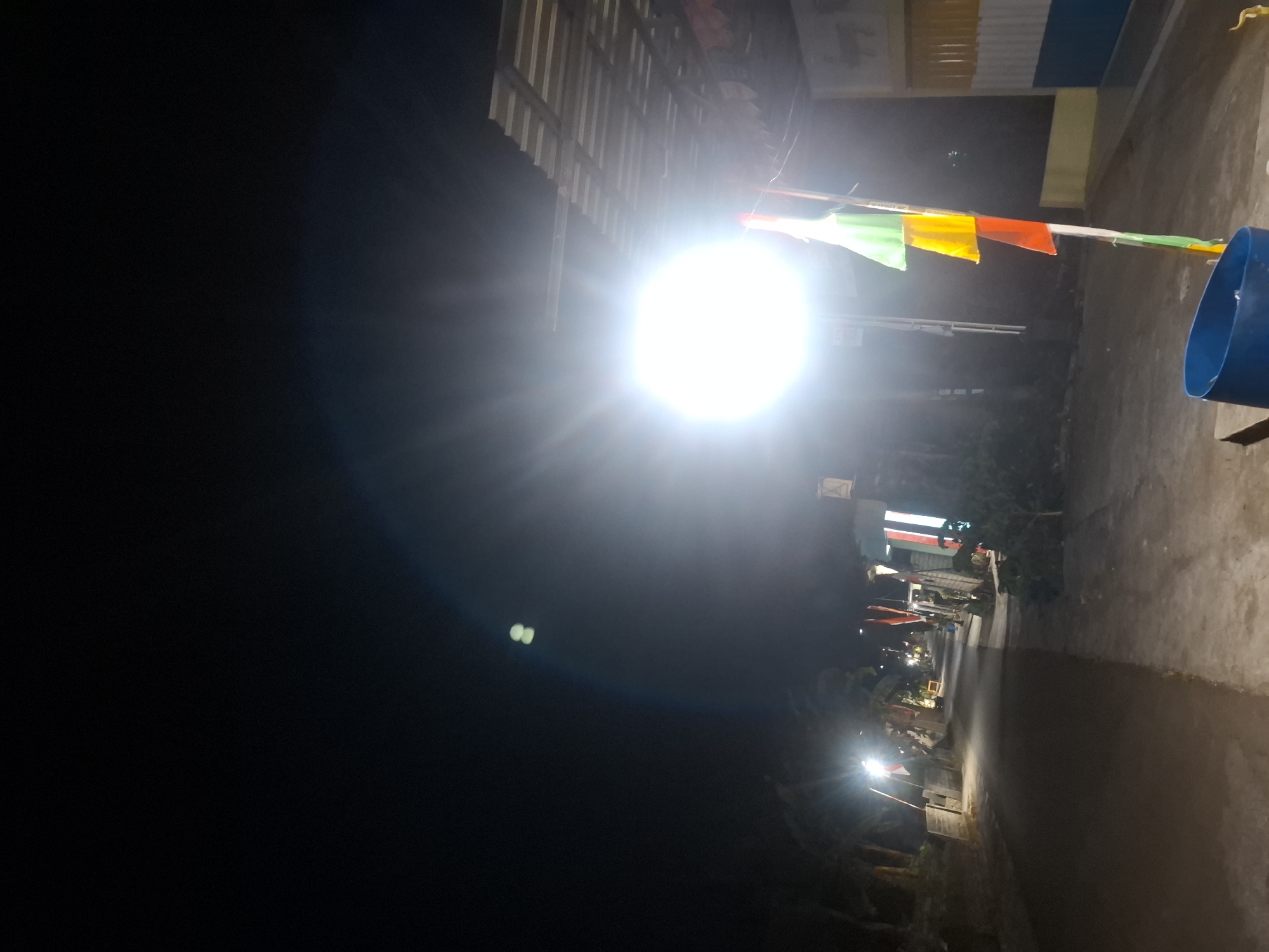 The image depicts a nighttime scene on a street in Bulubrangsi, Indonesia. A bright, artificial light source illuminates the area from the right side of the frame, creating lens flare and washing out details. On the right, a building with a corrugated metal roof is visible, with colorful flags attached to a pole in the foreground. The street itself appears to be paved, with a darker asphalt surface and a lighter concrete sidewalk. To the left of the street, trees and vegetation are discernible in the dim light. Several other light sources are present along the street, suggesting it is a populated area. The overall atmosphere is dark and somewhat obscured by the intense light source.