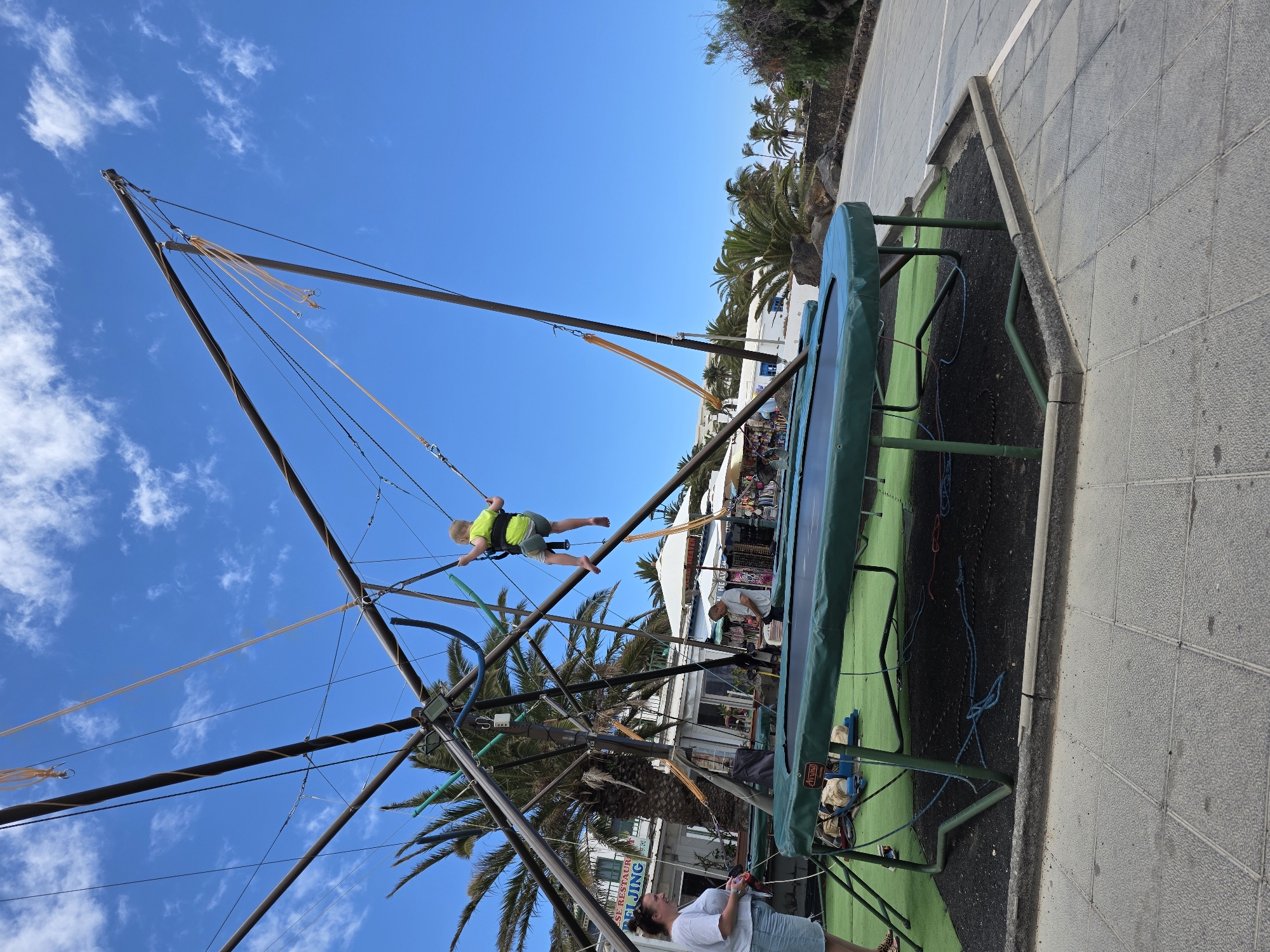 A young child in a neon yellow vest is mid-air, suspended by bungee cords attached to a large trampoline apparatus. The child is wearing a harness, and the bungee cords extend upwards to a central pole structure. The trampoline itself is green and has a metal frame. The scene appears to be outdoors on a sunny day, with a bright blue sky and scattered white clouds.
In the background, there are palm trees and buildings with white facades and blue accents, suggesting a warm, possibly coastal, location. Several people are visible near the trampoline; one adult appears to be operating the equipment or supervising, and others are observing. The ground around the trampoline is a mix of dark material and paved grey tiles. There is some text visible on the side of the trampoline, though it is partially obscured and difficult to read. The overall atmosphere suggests a recreational activity in a tourist-friendly environment. The time of day is likely daytime, given the bright sunlight and clear sky.