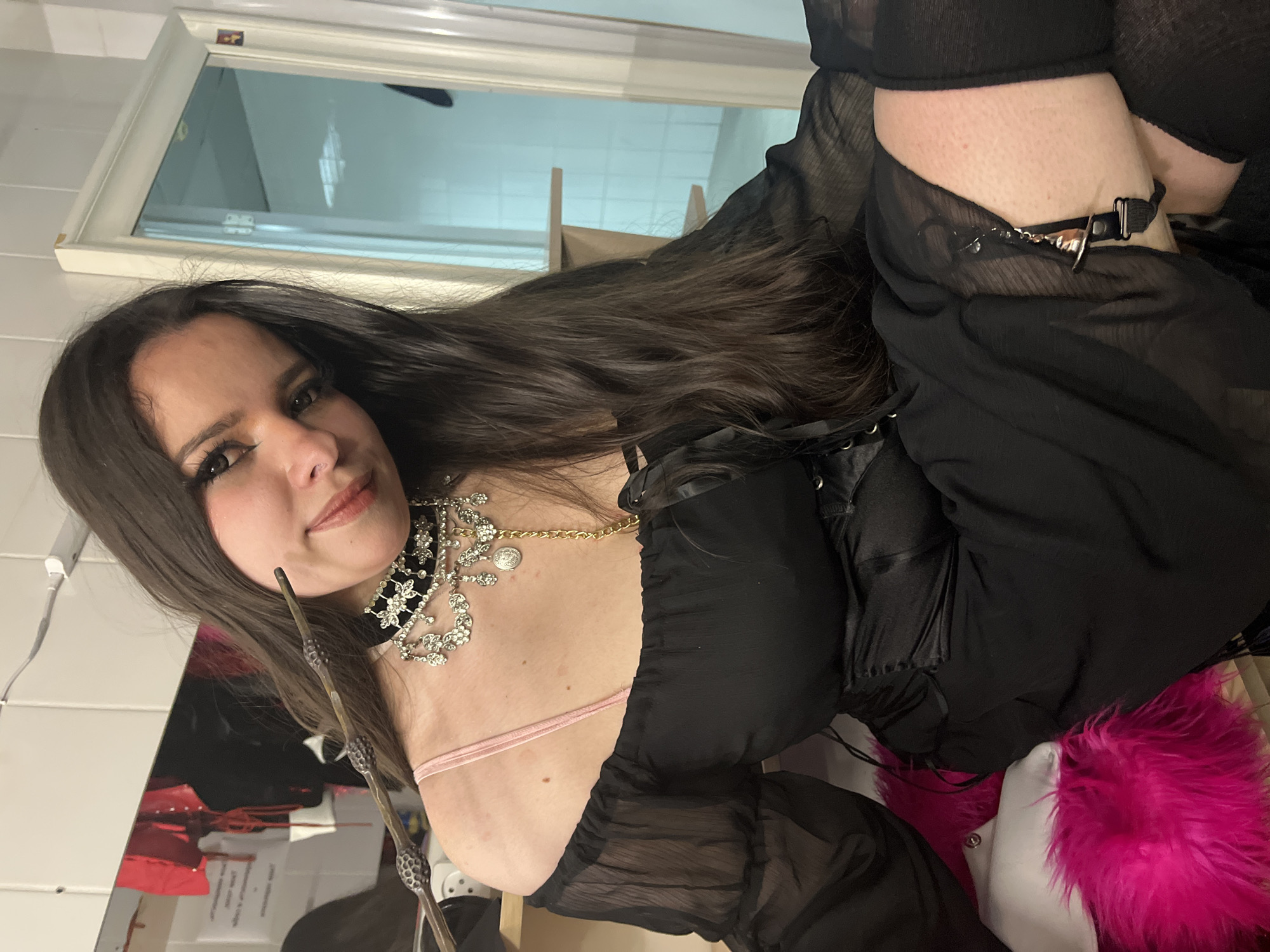 An indoor, eye-level shot depicts a female individual with long dark brown hair, seated and looking directly at the camera with a slight smile. She has dark eye makeup and is wearing a black off-the-shoulder top with sheer, puffed sleeves, revealing a pink bra strap on her right shoulder. A black corset or cincher is worn at her waist. She wears an elaborate silver-toned choker necklace with multiple hanging chains, clear stones, and a small gold-toned chain with a medallion. Her right hand holds a dark brown, textured wand-like object, pointed towards her left ear. Black thigh-high stockings are visible on her lower body, with a silver and black garter clip fastened to the stocking on her right thigh.
The background consists of white tiled walls. To the left, a large mirror reflects red garments, possibly lingerie, hanging on a rack, and a blurred dark figure. An electrical outlet is visible on the wall above the mirror. To the right, a white door frame with a glass panel reflects interior lights. A light-colored wooden shelf extends from the right wall. A vibrant pink furry object and a white cushion are partially visible in the lower-left foreground, where the individual is seated. The location is an indoor setting in the city of Unknown, Unknown.