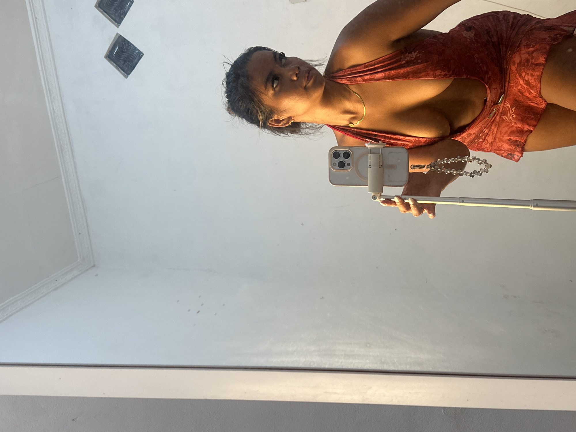 A woman is seen in a mirror selfie, holding a white phone with a phone mount on a selfie stick. The woman has brown hair and is wearing a low-cut, red dress with a gold necklace. The image is taken at an angle, showcasing the reflection in a mirror. The background shows a ceiling with two square vents. This is likely inside a home or building. The location, Celuk, Indonesia, is provided context.