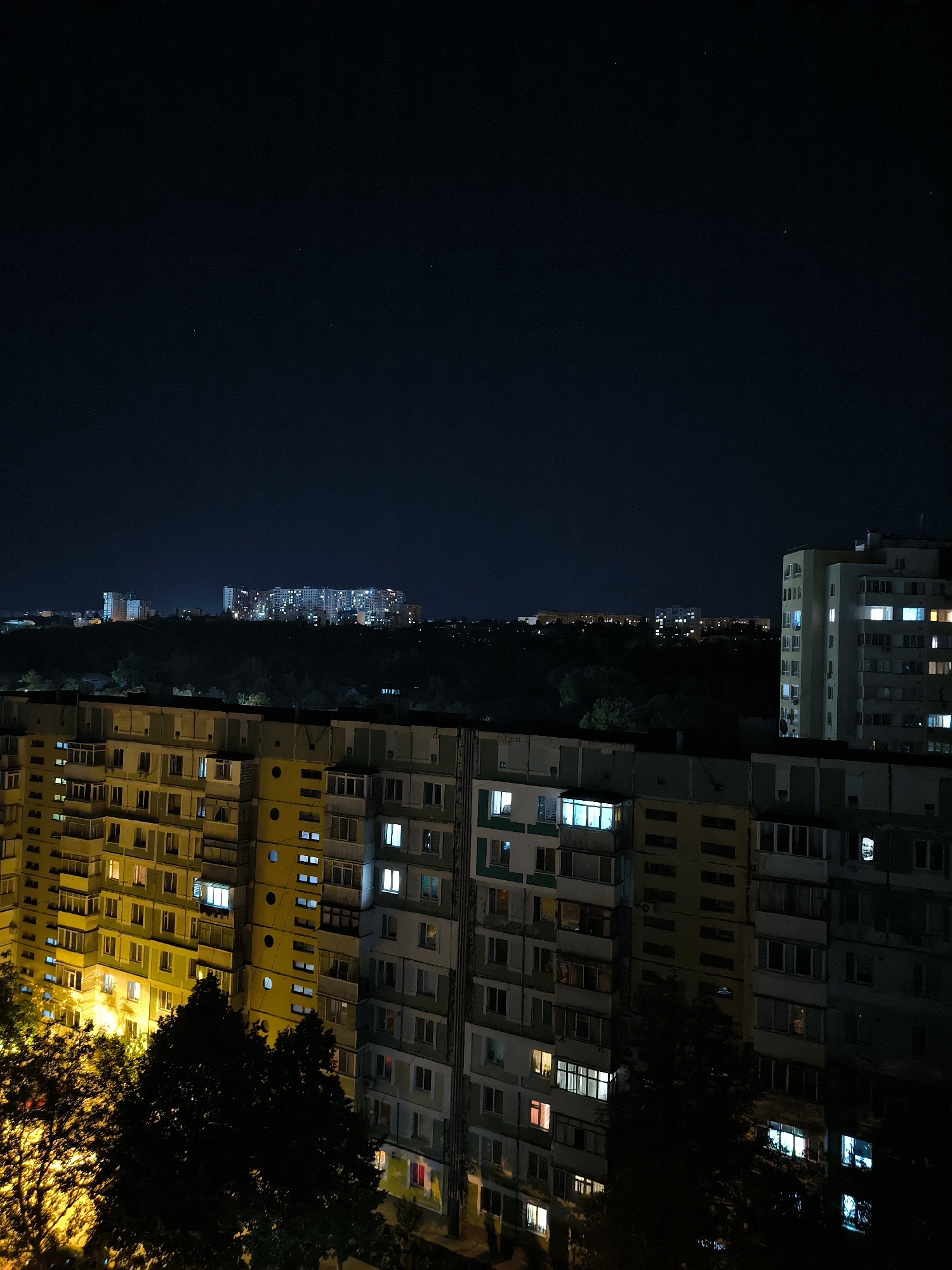 The image captures a nighttime view of a residential area in Chișinău, Moldova. In the foreground, a cluster of multi-story apartment buildings dominates the scene. These buildings, likely constructed during the Soviet era, exhibit a repetitive architectural style with numerous balconies and windows, many of which are illuminated, indicating occupancy. The facades appear weathered and utilitarian, with some sections painted in muted yellows and grays, while others are a lighter, possibly teal or pale green.
Beyond the immediate foreground buildings, a dark expanse of trees forms a dense line, suggesting a park or a green belt within the urban landscape. Further in the distance, a larger complex of apartment buildings is visible, also brightly lit with countless windows. These distant buildings are taller and appear more modern than those in the foreground, contributing to the layered urban panorama. The sky above is a deep, dark blue, characteristic of a clear night, with a few faint stars visible. The overall impression is one of a typical urban night in a residential district, characterized by the interplay of artificial light and darkness.