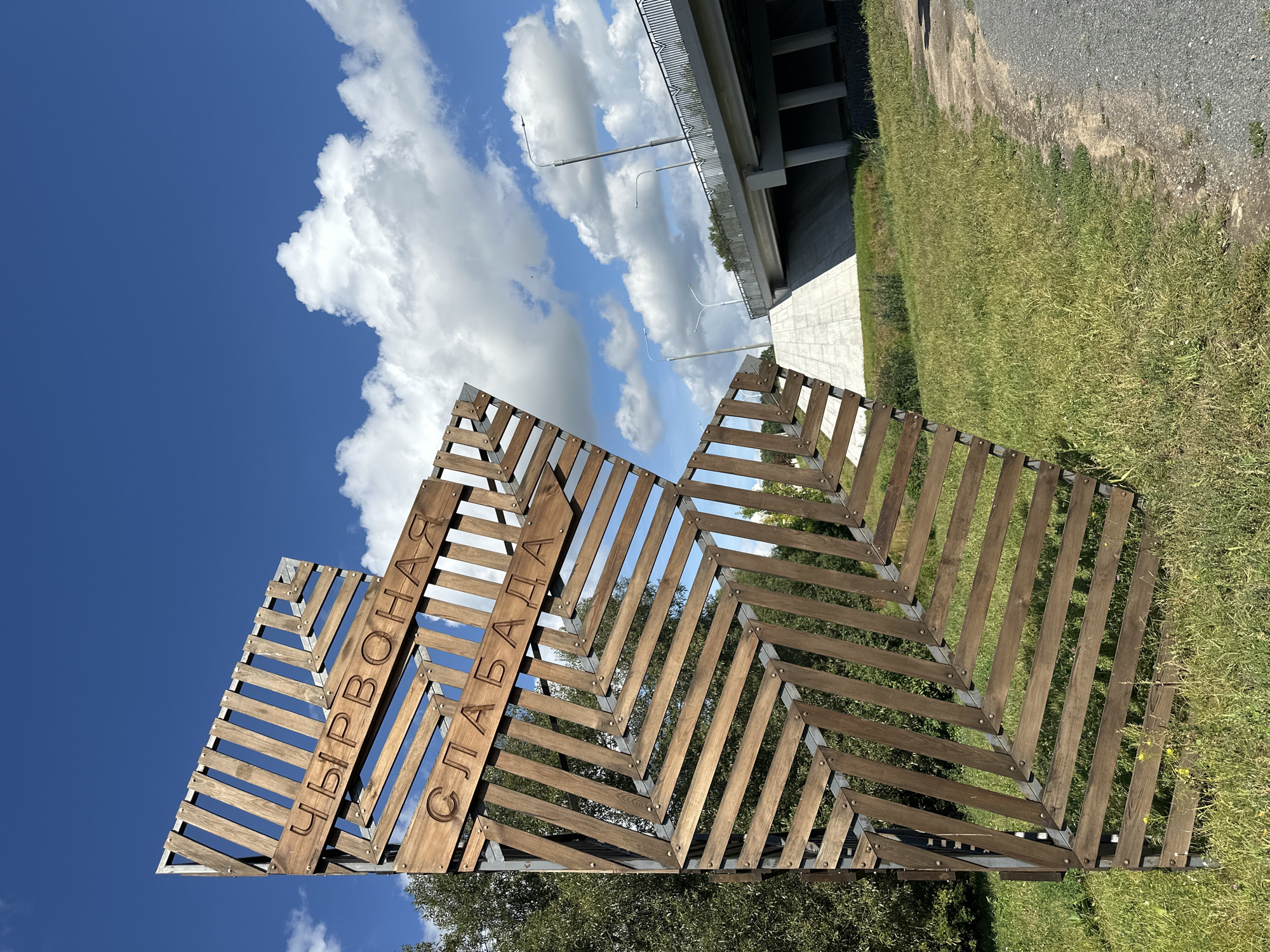 The image captures a wooden art installation with Cyrillic lettering that reads "ЧЫРВОНАЯ СЛАБАДА" (Chyrvonaya Slabada), which translates to "Red Sloboda." This installation is situated outdoors, with a bright blue sky and scattered white clouds serving as the backdrop. To the right of the installation, a modern concrete bridge with railings and streetlights spans across the scene, indicating an urban or suburban environment. The ground is covered in green grass and a gravel path runs along the right edge of the image. The overall impression is that of a public space, possibly a park or a roadside attraction, in Minsk, Belarus, as indicated by the "Red Sloboda" street name which is a known location in the city. No people are visible in the image.
