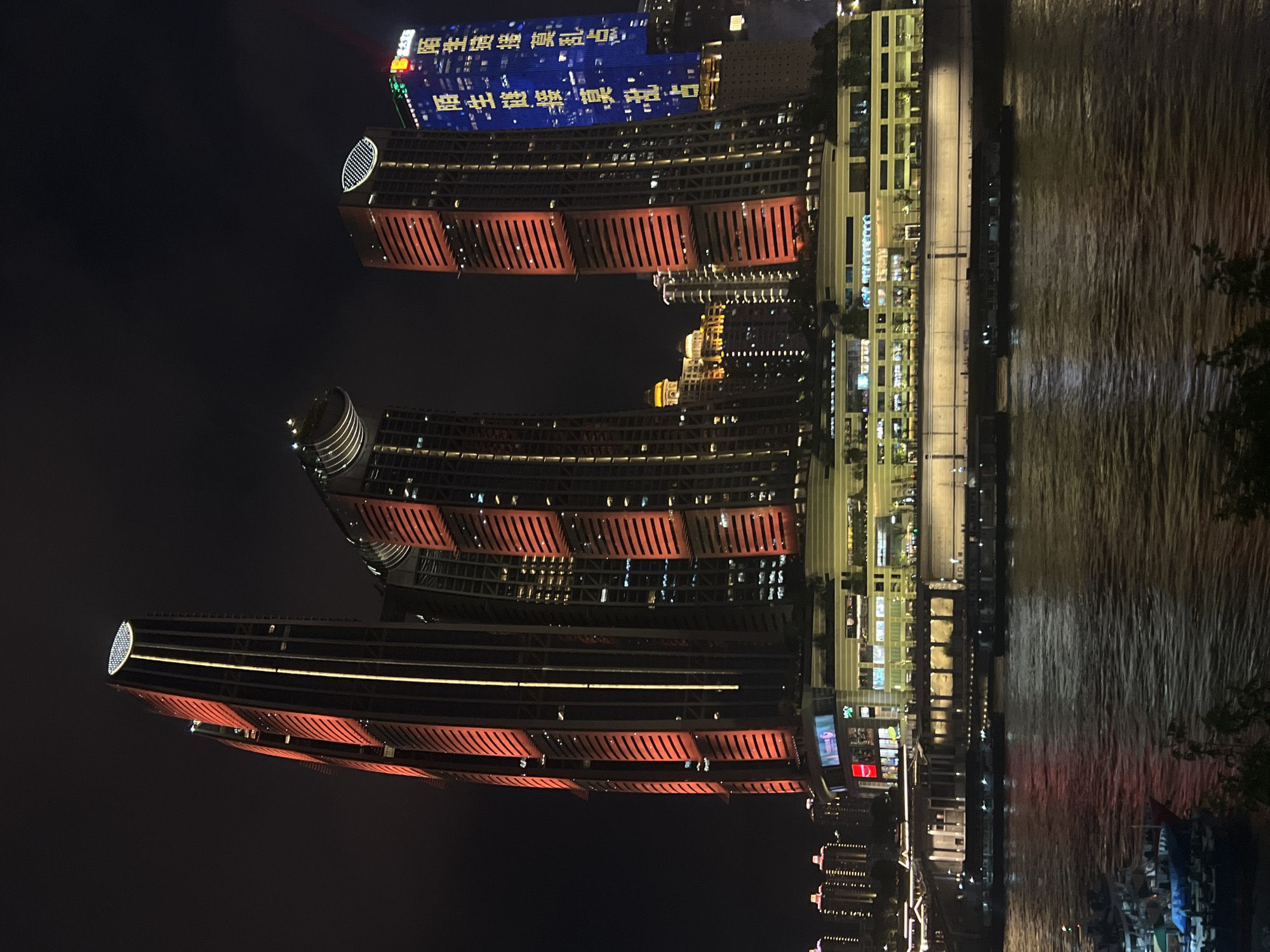 The image depicts the Chongqing Rail Transit Lines 6 and 9 station, also known as the Jiangbeicheng Street, China, at night. The iconic building features a complex of three interconnected skyscrapers, with the tallest on the left displaying a prominent oval-shaped LED display at its apex. The buildings are illuminated with red and yellow lights, creating a striking contrast against the dark sky. In the foreground, a wide river flows, reflecting the city lights. Several boats are visible on the water, with one large vessel docked on the left side of the frame. The background shows a cluster of residential buildings also lit up, adding to the urban landscape. The scene is characterized by its modern architecture and vibrant nighttime illumination.