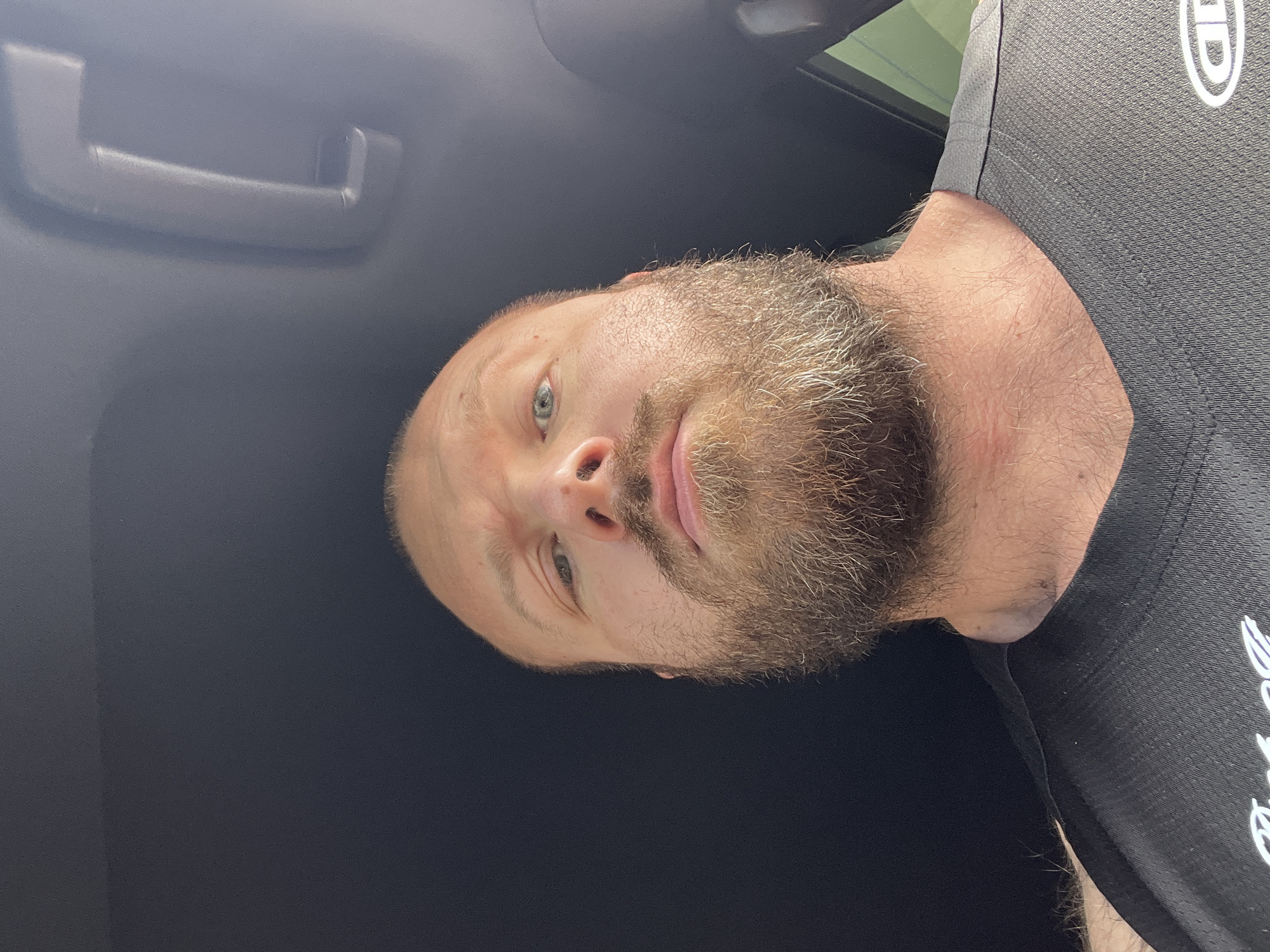 A bald, bearded Caucasian male, estimated to be in his 30s or 40s, is captured in a close-up, low-angle selfie. He has a full, reddish-brown beard and mustache, light-colored eyes (appearing blue or green), and a fair complexion. A small mark is visible on the bridge of his nose. He is wearing a dark, possibly black or dark grey, short-sleeved athletic shirt with a V-neck design. A partial white logo, resembling an oval with text, is visible on the right side of his chest. The man is looking directly into the camera with a neutral expression. The background indicates he is inside a vehicle, with a dark headliner and a grey overhead grab handle visible in the upper right quadrant of the image.