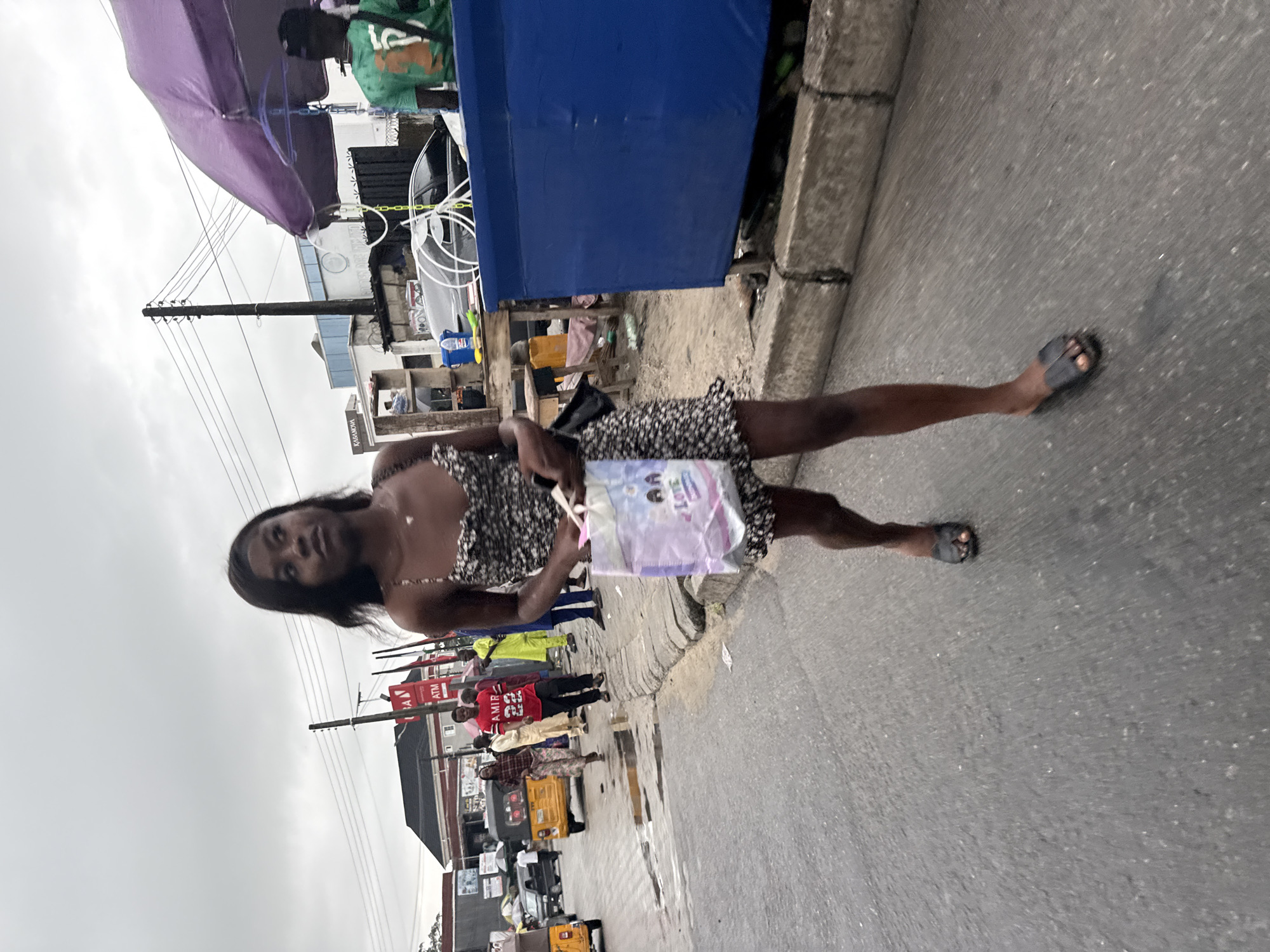 In Ado, Nigeria, a young Black woman walks on a paved road with a gray, overcast sky. She is wearing a black and white floral print dress and dark sandals, and she carries a white shopping bag with colorful graphics, including what appears to be cartoon characters and the word "LOVE". In the background, other individuals are visible, including a man in a red shirt with "AMIR 22" printed on it, another man in a bright green shirt, and people near a yellow auto-rickshaw. Buildings with signage and utility poles are also present, suggesting an urban environment. The road shows signs of wear and puddles, indicating recent rain. A blue stall with a purple umbrella is situated on the right side of the frame.