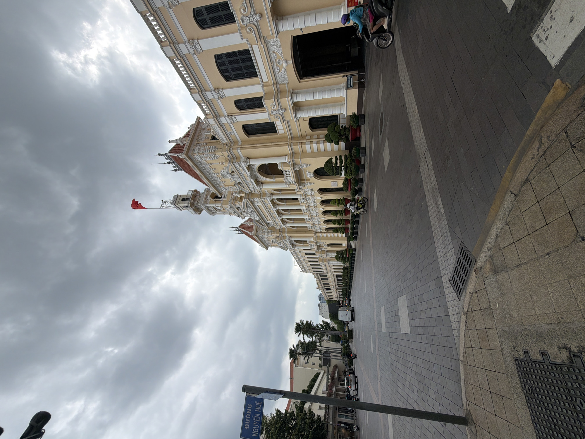 This image depicts the Ho Chi Minh City Hall (Trụ sở Ủy ban Nhân dân Thành phố Hồ Chí Minh) situated on Nguyễn Huệ Street in Ho Chi Minh City, Vietnam. The grand, multi-story building occupies the right side of the frame, showcasing a pale yellow facade with ornate white architectural detailing, including columns, balconies, and cornices. A central clock tower, topped with a red Vietnamese flag, rises above the building. Dark-shuttered windows and arched entryways are visible along its length, with manicured potted plants lining the base.
The foreground features a wide, paved pedestrian street, identified by a blue sign on the left as "ĐƯỜNG NGUYỄN HUỆ" (Nguyễn Huệ Street). The street surface consists of varying shades of gray and white interlocking pavers, with metal grates visible on the ground. On the far left, a dark pole stands prominently. Further down the street on the left, other urban buildings and several palm trees are visible.
Sparse traffic includes a white car parked on the left, a white van driving in the mid-ground, and multiple motorcycles. One person on a black scooter, wearing a blue top, patterned shorts, and a purple helmet, is positioned on the right side of the street near the building. Another person on a scooter is visible further down the street on the right.
The sky is overcast with thick, gray clouds, casting a soft, diffused light across the scene. The overall environment is an urban streetscape with grand architecture.