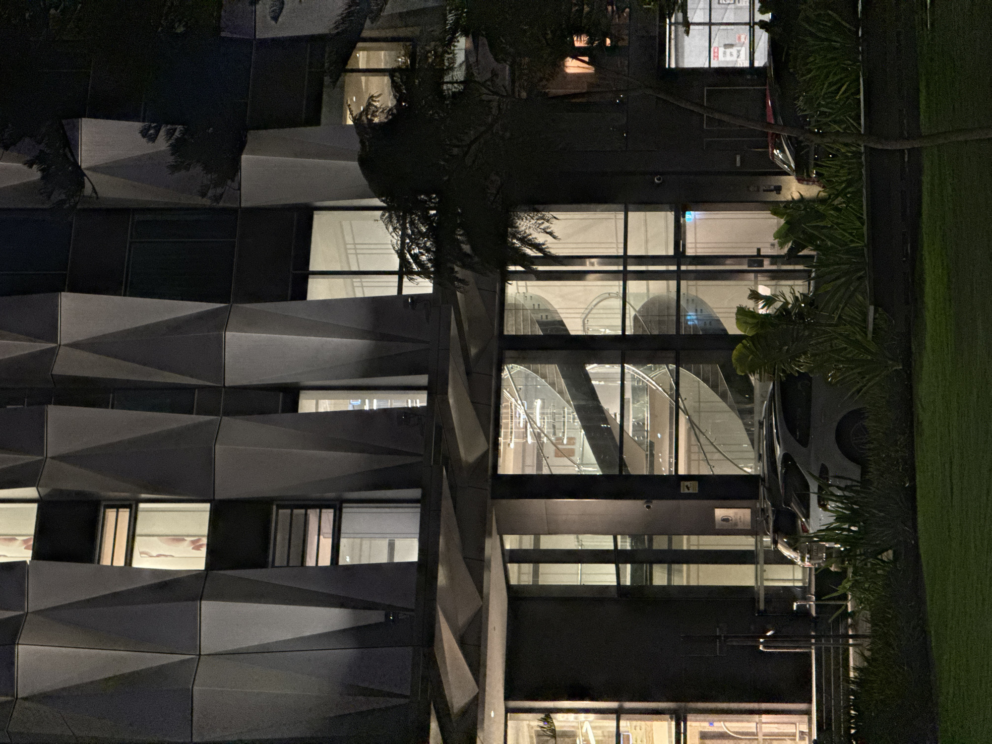 The media file presents a vertical orientation image, depicting the exterior of a modern building at night in Perth, Australia. The structure dominates the left and center of the frame, characterized by a dark, geometric facade composed of numerous angled, faceted panels, possibly made of metal or concrete. Interspersed within this striking design are various rectangular windows. Some windows on the upper levels glow with interior light, revealing glimpses of indoor spaces, while larger glass sections on the right side of the building clearly display an illuminated internal structure, resembling an escalator or a curving staircase.
To the right of the building, a strip of vibrant green grass runs vertically, indicating a landscaped area. Lush green bushes, possibly palms or other tropical foliage, are situated between the building and the grass, partially obscuring the lower sections of the facade. A dark, leafy tree with intricate branches stands in the mid-ground, its silhouette cutting across the building's facade. A light-colored car is partially visible, parked behind some bushes on the far right side of the image, near the edge of the grass.
The scene is static, with no discernible human activity or movement. The time of day is clearly night, as evidenced by the dark sky and the artificial illumination emanating from within the building. The weather appears calm. Notable details include the distinct architectural style of the building, which lends a contemporary and possibly commercial aesthetic. While there are small signs visible on the building's exterior and through windows, the text on them is too blurry to be deciphered.