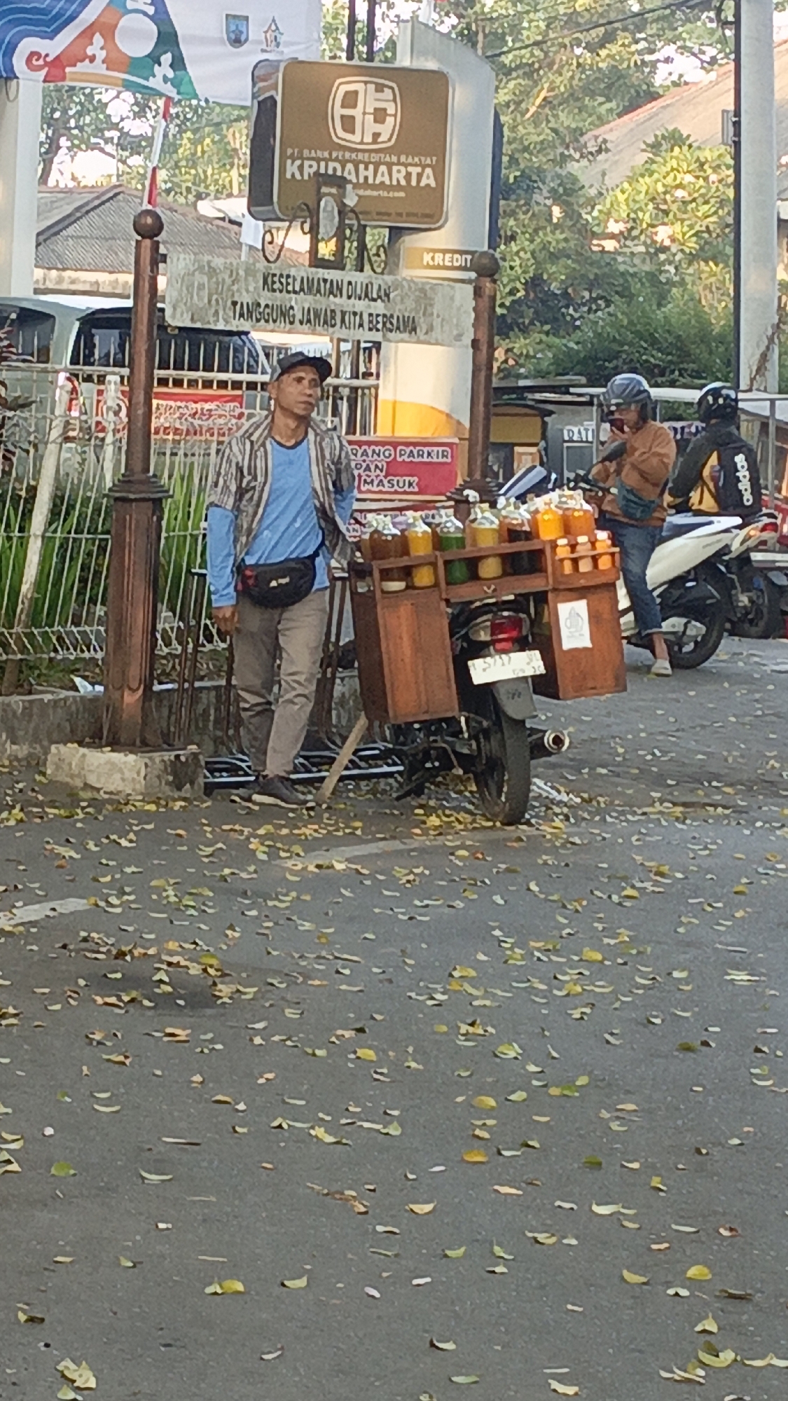 A street vendor stands next to a motorcycle modified to carry a wooden box filled with bottles of various colored liquids. The vendor, a man wearing a blue long-sleeved shirt, a striped open jacket, and gray pants, has a black fanny pack around his waist and a dark cap on his head. He is looking upwards and to his left. The motorcycle, parked on the side of the road, has a license plate that reads "F 5717 JD". Behind the vendor, a sign reads "KESELAMATAN DIJALAN TANGGUNG JAWAB KITA BERSAMA" (Road Safety is Our Shared Responsibility). Above this sign, another sign displays "PT. BANK PERKREDITAN RAKYAT KRIDAHARTA". The ground is littered with fallen leaves, suggesting an outdoor, possibly urban environment. The presence of motorcycles and the style of the signs indicate this is likely in Indonesia, specifically the city of Salatiga, which is known for its bank and the general context.