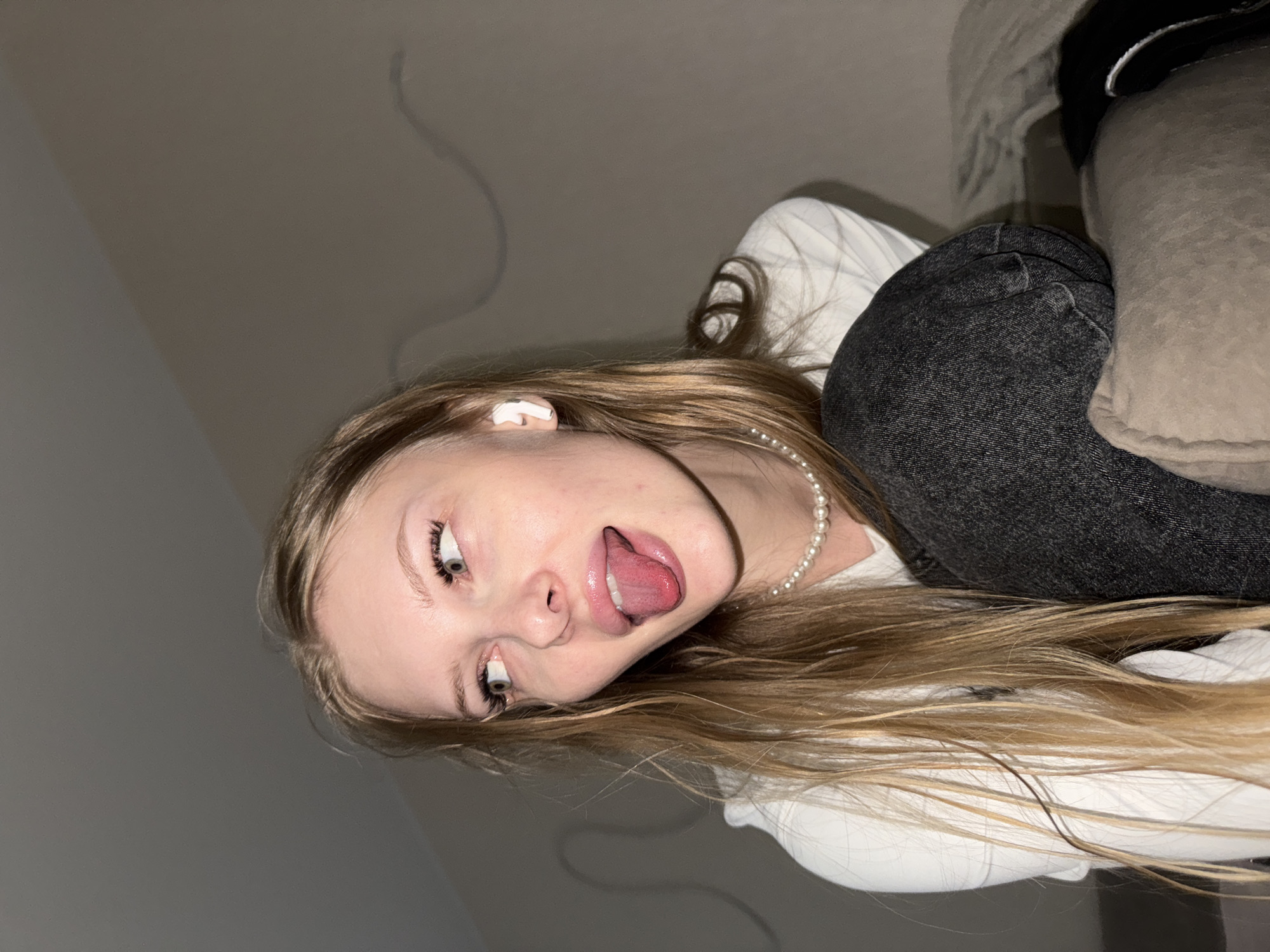 An informal, close-up portrait taken indoors in Perm, Russia, shows a young individual, likely female, with long, straight blonde hair, fair skin, and light-colored eyes. The image is oriented sideways, with the subject's head tilted.
She is wearing a white long-sleeved top and dark, possibly black or gray, jeans. A delicate pearl necklace adorns her neck, and a white earbud is visible in her left ear. Her expression is playful and exaggerated; she has her eyes rolled back and her tongue sticking out.
The background consists of a plain, light gray wall, with a faint, dark, curved line, possibly a cable or its shadow, visible on the surface. To her right, a soft, light-colored fabric object, such as a pillow or blanket, is partially visible. The lighting appears soft and diffused, characteristic of indoor artificial light. The overall mood is casual and humorous. No text is visible in the image.