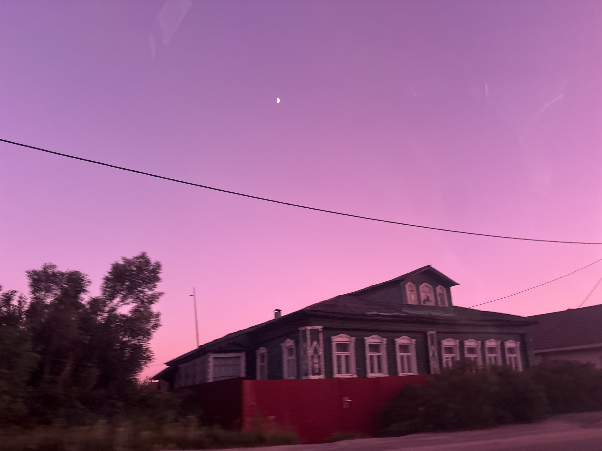 An eye-level, slightly unfocused, medium-shot photograph depicts a two-story, dark-green house with white-trimmed windows under a pink sky in Zaprudnya, Russia. The house has a red lower wall or fence. To the left of the house is a tree; to the right is a neighboring house. A thin power line bisects the frame horizontally. A crescent moon is visible in the upper left.