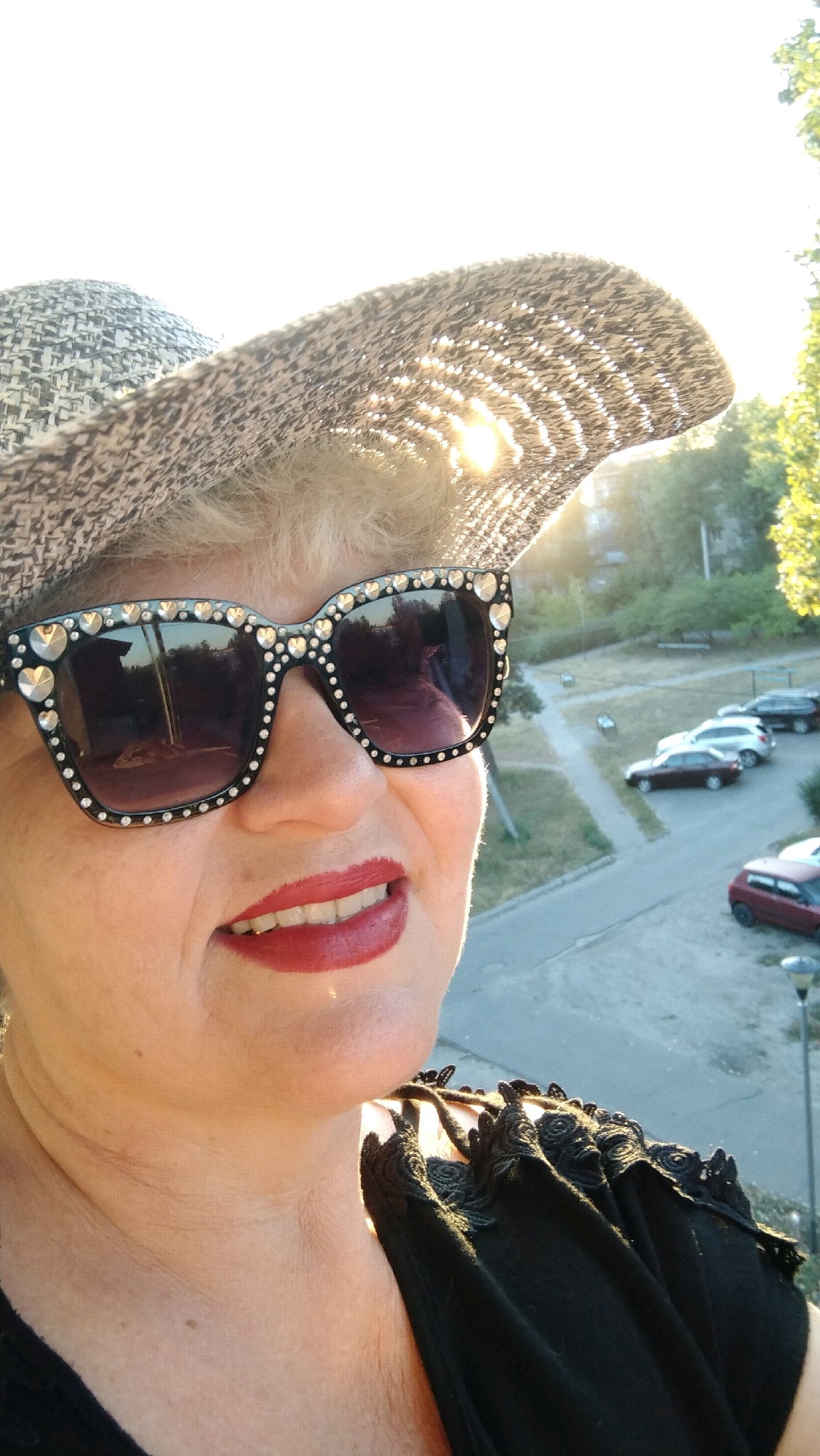 A woman wearing a wide-brimmed hat and large, jeweled sunglasses smiles at the camera. She has bright red lipstick on. The hat is adorned with a black and white patterned band and is casting a shadow over her head. The sun is visible through the weave of the hat. In the background, there is a view of a city street with parked cars and trees. The image appears to be taken from a higher vantage point, possibly a balcony or window, in Horishni Plavni, Ukraine. The woman is wearing a black top with lace detailing on the shoulder.