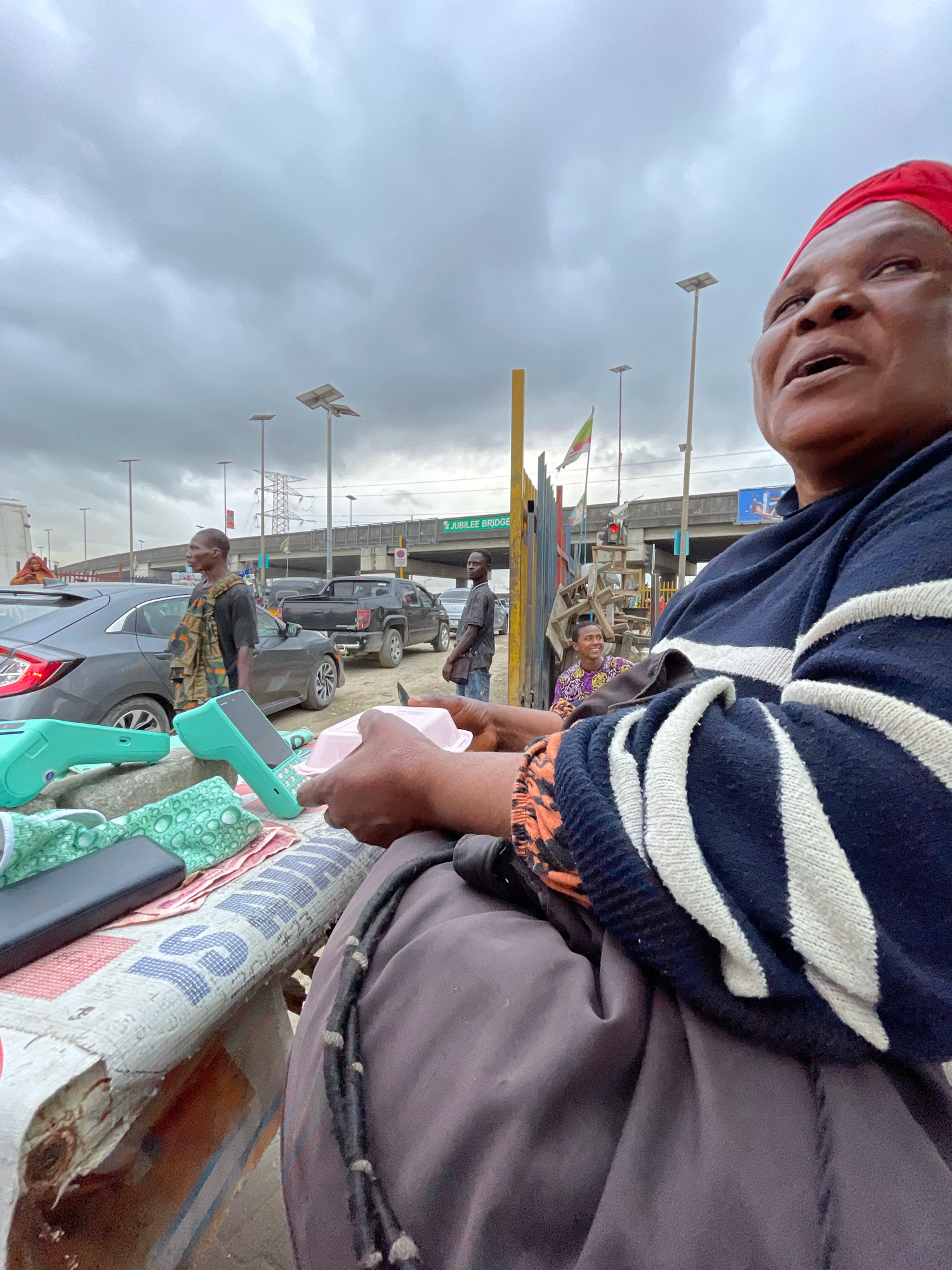 A woman wearing a red headscarf and a blue and white striped sweater is sitting on a stool at a street market in Ajah, Nigeria. She is looking to the side, with her mouth slightly open. In front of her are several items laid out on a table, including two mint green point-of-sale devices and a phone. Behind her, a busy street scene unfolds. Cars are driving by, and other people are walking around. A bridge with the sign "JUBILEE BRIDGE" is visible in the background, along with streetlights. The sky is overcast with gray clouds. The overall atmosphere is one of everyday urban life in a market setting.