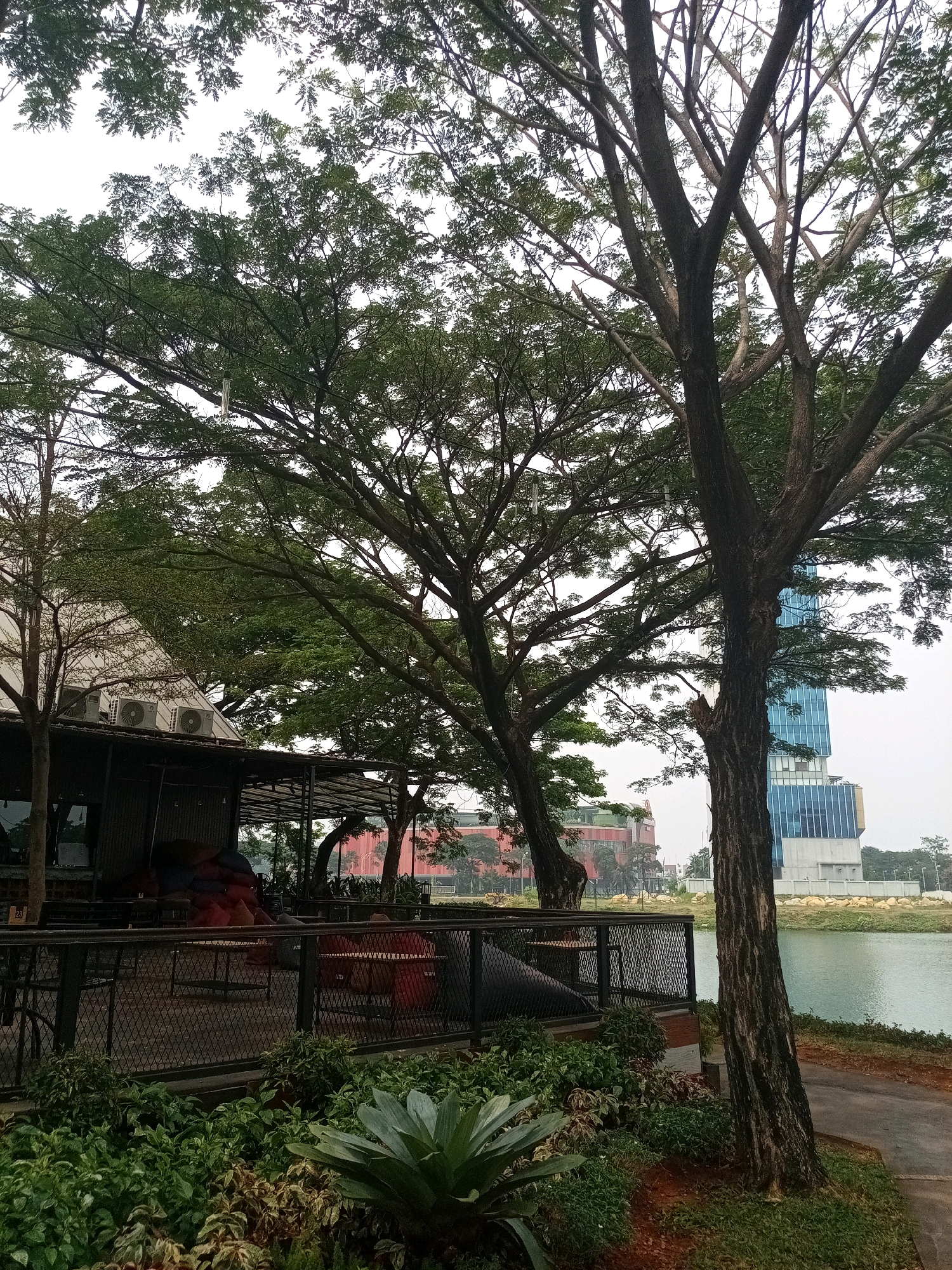 The image shows an outdoor seating area of a restaurant or cafe located in the city of Bekasi, Indonesia, adjacent to a body of water. In the foreground, there are lush green plants, including a large agave-like plant. Behind the plants is a dark-colored metal fence surrounding an outdoor seating area with tables and chairs. Some colorful cushions are visible on some of the seating. Above the seating area, there is a building with a white, angled roof and several outdoor air conditioning units mounted on it. In the background, several large trees with dense green foliage are prominent, with a tall, modern glass-fronted building visible behind them and to the right. The body of water in the background appears to be a lake or large pond. The sky is overcast.