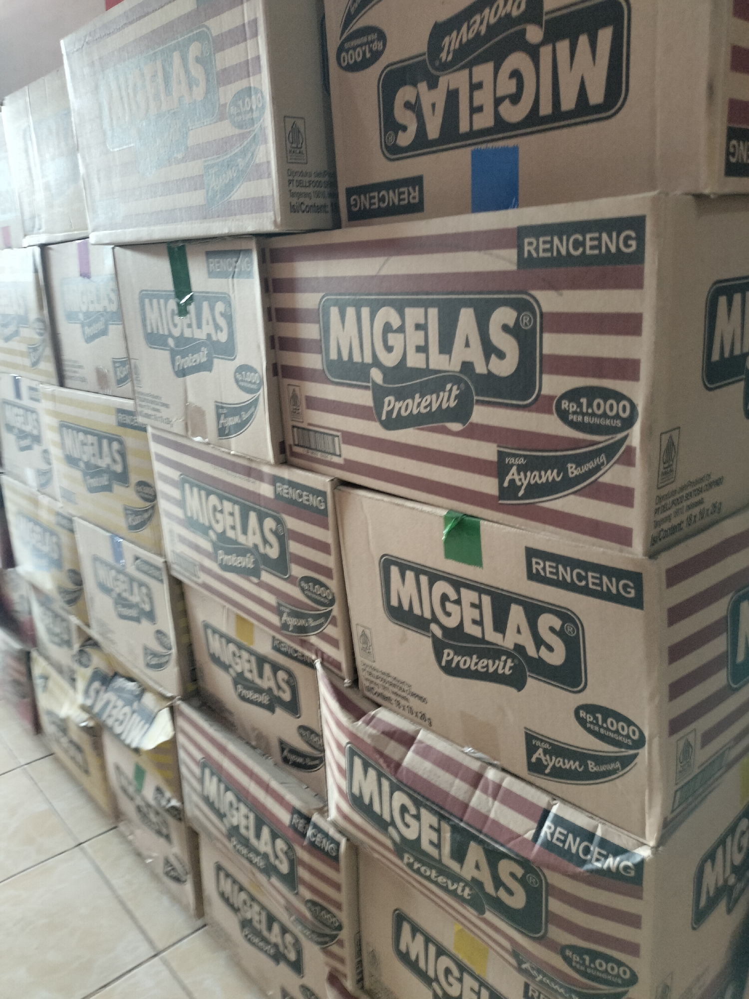 The image shows a large stack of cardboard boxes, predominantly featuring the "MIGELAS Protevit" brand. The boxes are stacked in multiple rows and columns, suggesting a retail or storage environment. Most of the boxes are oriented so that their fronts, displaying the product name and branding, are visible. The color palette of the boxes is primarily light brown with blue and red accents. Some boxes also show pricing information, such as "Rp.1.000 PER BUNGKUS". In the background, additional boxes and a wall can be observed. The floor appears to be tiled. The lighting suggests an indoor setting, possibly a warehouse or a shop aisle. The image does not depict any people, actions, or interactions. The context of Langsa, Indonesia, is mentioned, but there are no specific visual cues in the image that confirm this location.