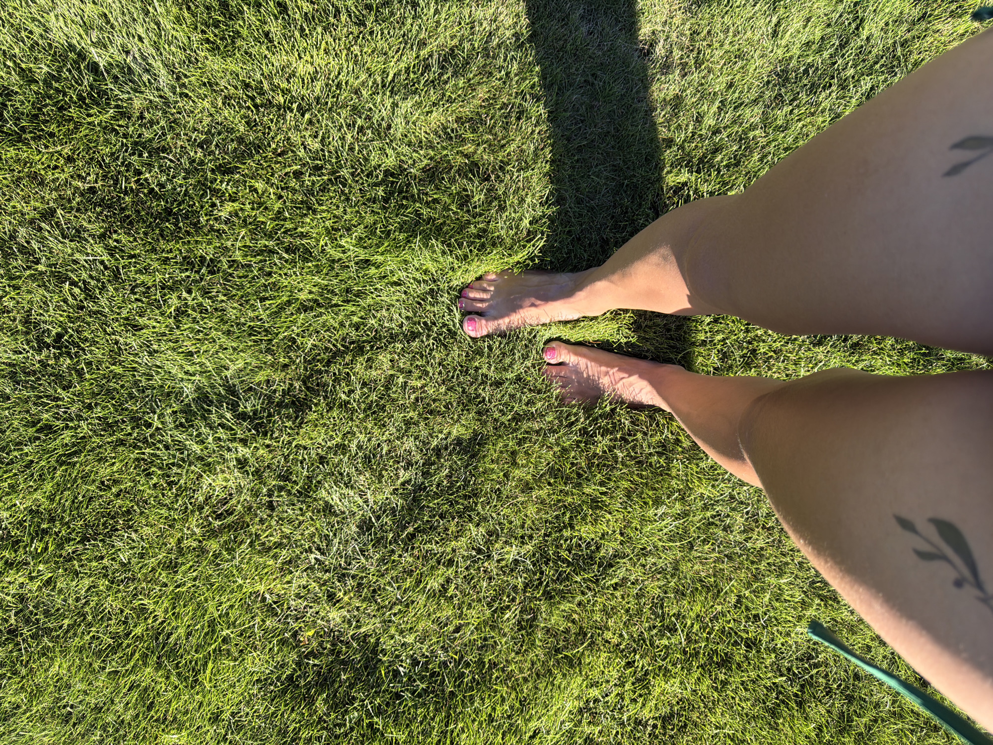 A top-down, medium shot shows a person's bare legs and feet standing on bright green grass under direct sunlight. The person's right foot is closer to the camera and to the left of the frame, with toenails painted pink. The left foot is further away and to the right, angled slightly. The person has a small, dark leaf tattoo on their upper left thigh. A thin, green cord is visible on the lower left edge of the frame. The scene appears to be outdoors on a sunny day, with a distinct shadow cast across the grass from the right side of the frame. The image does not provide enough information to determine the specific location in Novosibirsk, Russia.