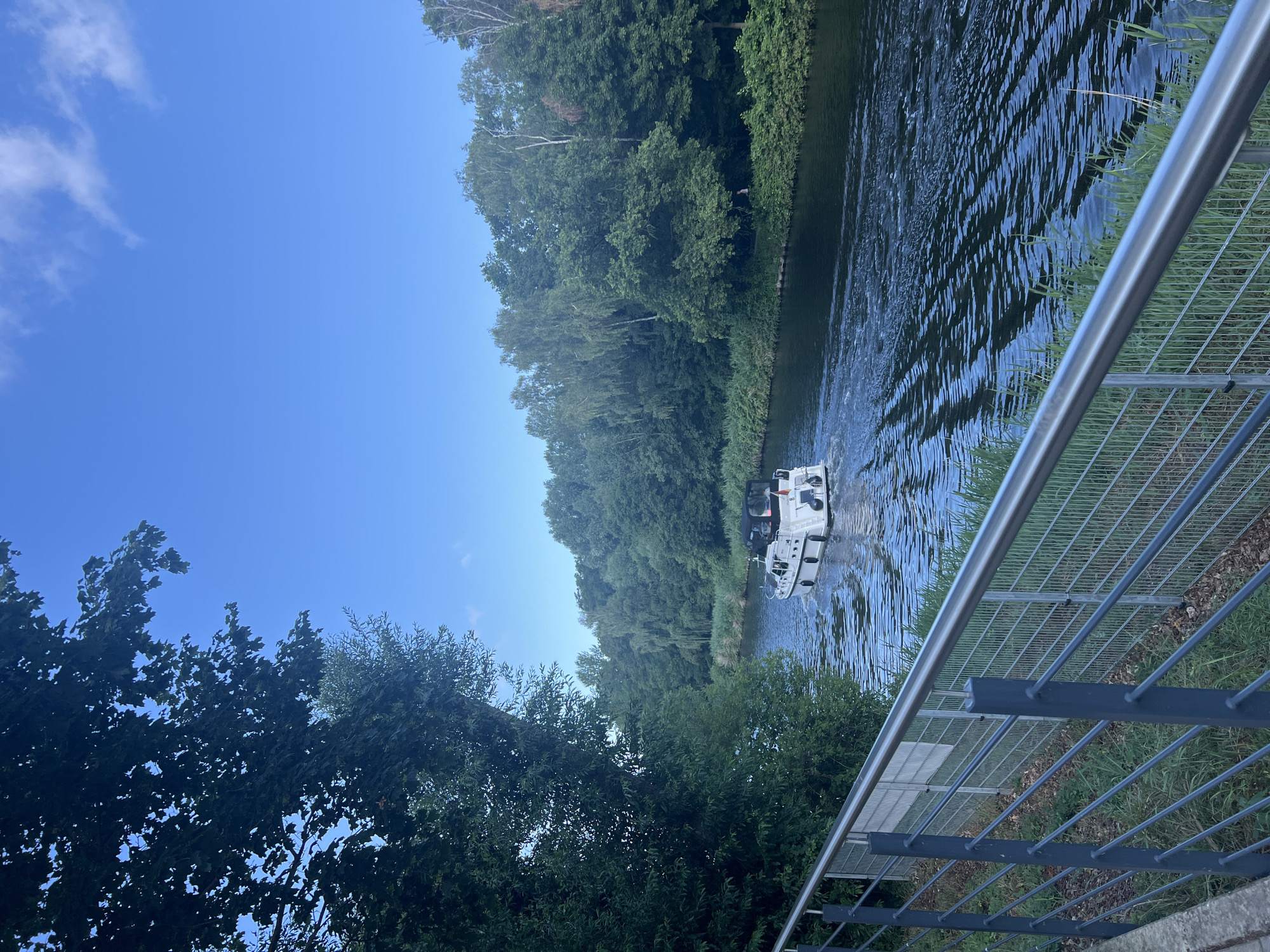 The image shows a body of water, likely a canal, with a white boat with a black canopy traveling away from the viewer. The boat is leaving a wake in the water. The canal is flanked by lush green trees on both sides, forming a dense forest. In the foreground, there is a metal railing and a concrete embankment, with some grass and fallen leaves. The sky is blue with a few scattered white clouds. The scene appears to be a peaceful waterway within a city, possibly Teltow, Germany, given the context. The overall impression is one of a serene, natural landscape within an urban setting.