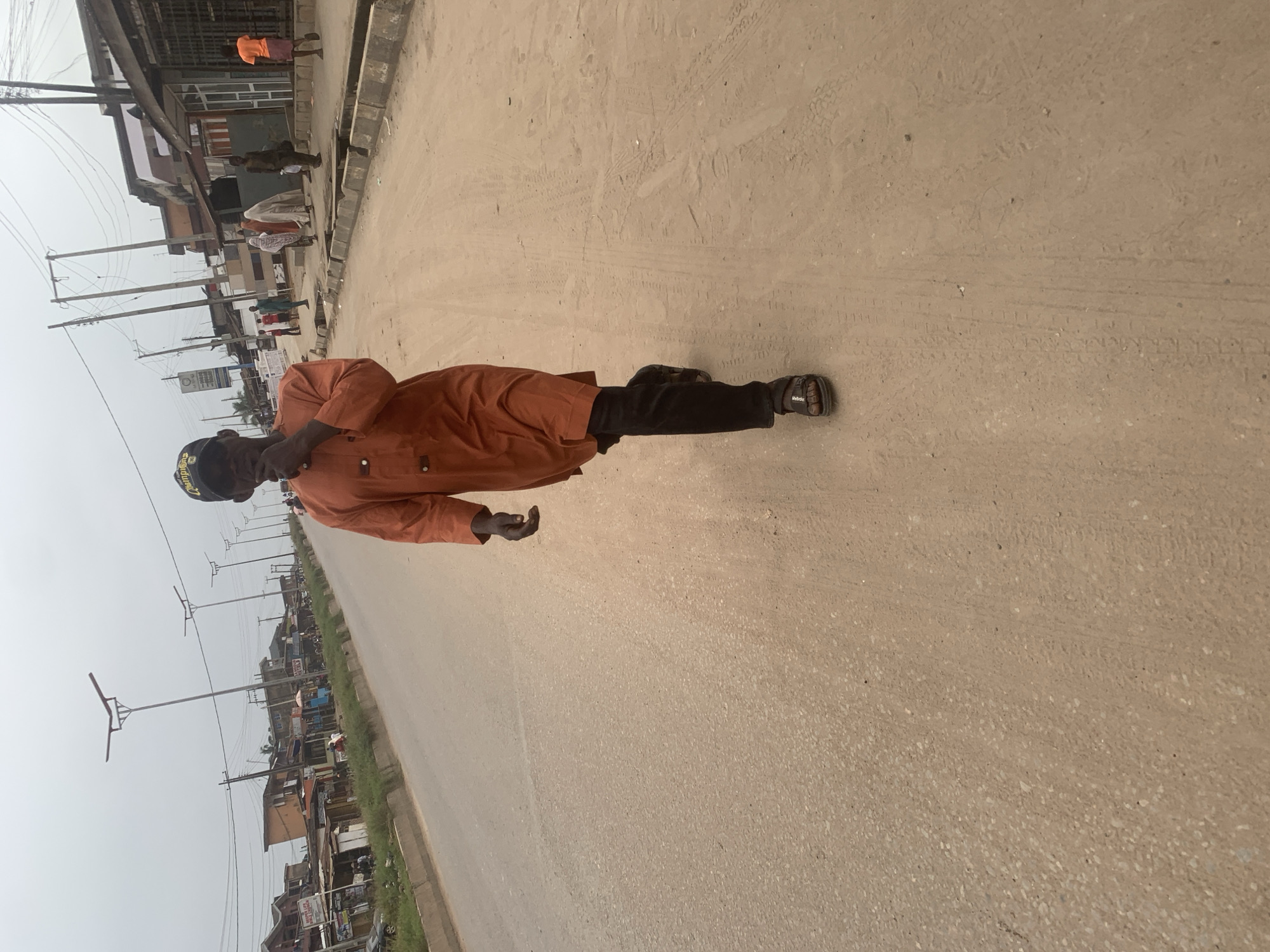 A man wearing an orange tunic and black pants walks on a dusty, paved road in Shagamu, Nigeria. He is wearing a black baseball cap and dark sandals. The man is holding his hand up to his face, possibly touching his mouth or nose. The road is lined with buildings on the right side of the image, appearing to be shops and residences. There are streetlights along the road. Several other people are visible in the background walking on the sidewalk or road, including a woman in a white veil and a child in an orange shirt. The sky is overcast.