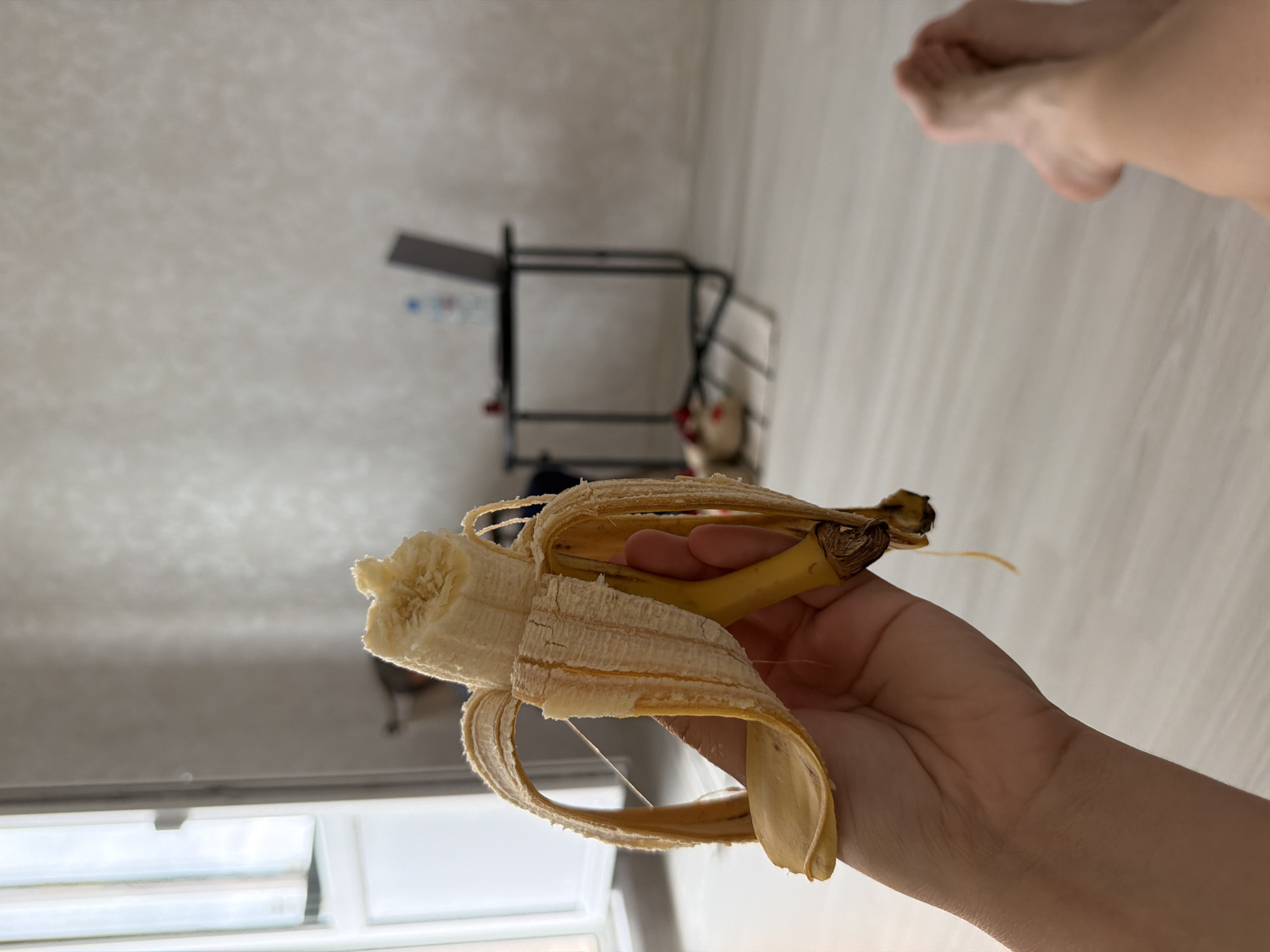 A person is holding a partially peeled banana in their hand. The banana is yellow with some brown spots, and the peel is hanging open. The person's hand is light-skinned. In the background, there is a room with light-colored walls and a light wooden floor. A small table with a laptop and a bottle of water is visible. A window with white curtains is on the left side of the image. A pair of feet are visible in the lower right corner of the frame. There is no discernible connection to the city of Froly, Russia.