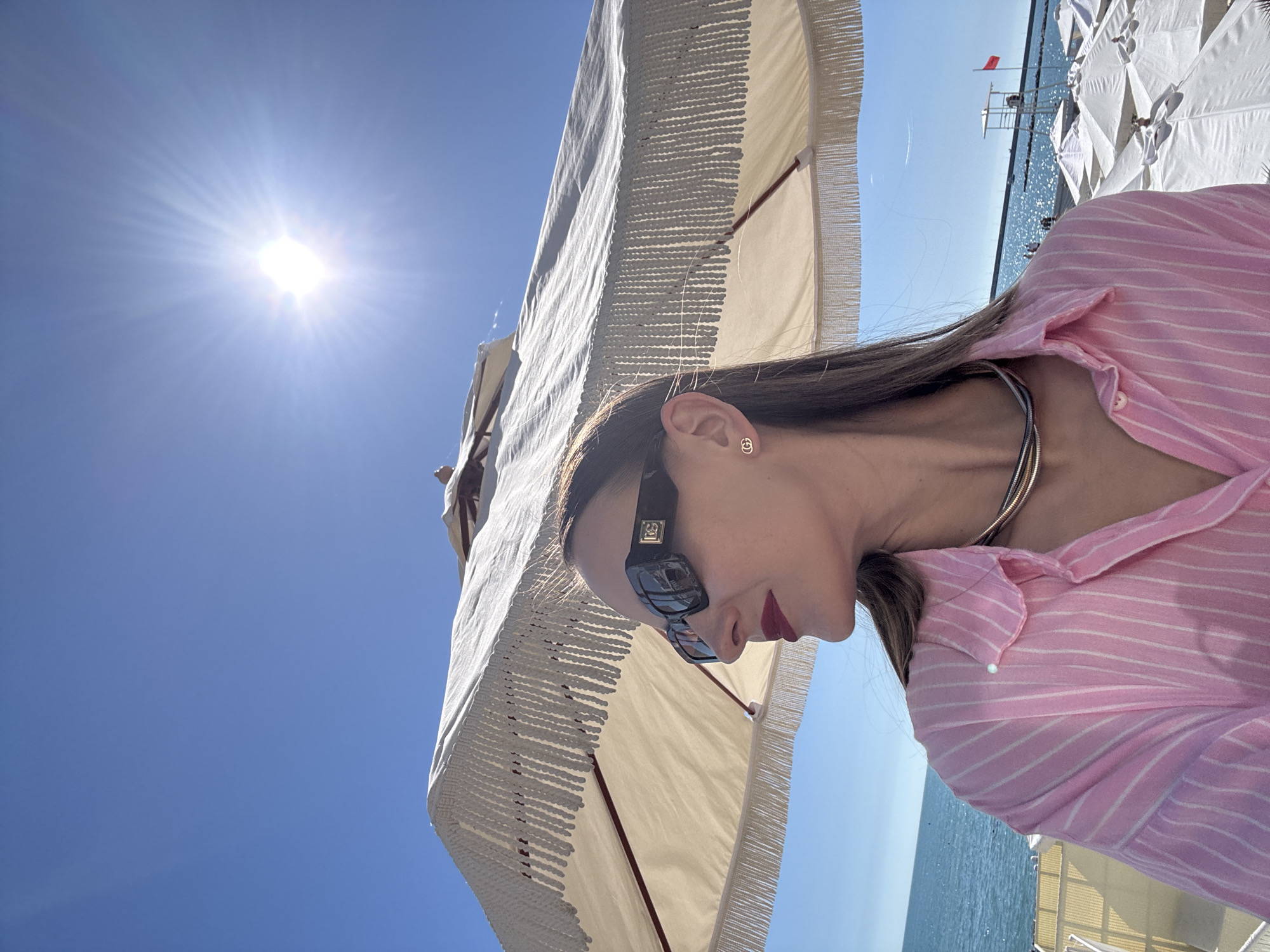 The image, captured from a low angle and rotated 90 degrees clockwise, depicts a sunny beach scene in Sochi, Russia. In the immediate foreground on the right, a person, likely female, is shown in profile from the chest up. She wears a light pink button-up shirt with thin white stripes and a multi-strand necklace, appearing to be silver and gold in color. Dark hair frames her face, and she is wearing dark-framed sunglasses, one arm of which displays a small, indistinct gold emblem. A small, golden stud earring, shaped like interlocking "G"s or a similar logo, is visible in her ear. Her lips are painted with dark red lipstick.
Above and behind her, a large, cream-colored beach umbrella with decorative fringe is partially open, providing shade. The sky is a vibrant, clear blue, dominated by a bright, intense sun in the upper left, casting a strong sunburst effect.
In the background, to the left, the sparkling blue sea extends towards the horizon, reflecting the sunlight. Several other white beach umbrellas are visible lining the shore, and a lifeguard stand, marked with a prominent red flag, stands out against the water. The scene conveys a relaxed and sunny day at the seaside. No distinct text is clearly visible.