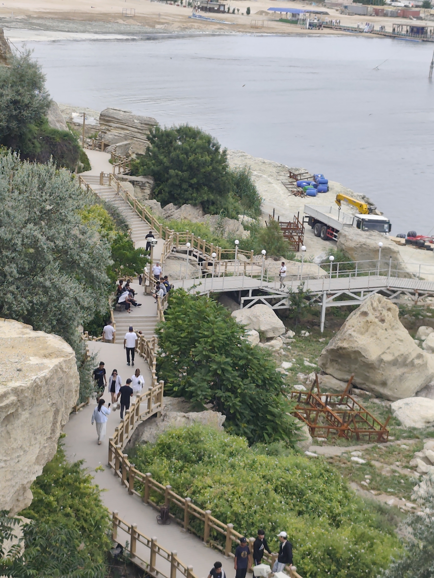 The image captures a scenic view of a coastal park area, likely in Aktau, Kazakhstan, with a body of water in the background. A winding paved pathway with wooden railings guides visitors along a rocky, elevated terrain adorned with lush green vegetation. Several people are seen strolling along the path and resting on benches, enjoying the outdoors.
In the distance, a sandy area transitions into the calm, grey water of a sea or large lake. Along the shoreline, there are signs of construction or development, including a yellow crane, a truck, and some blue barrels. A wooden walkway or bridge crosses over a lower section of the park, with people walking on it.
The lighting suggests it is daytime, possibly overcast, with no strong shadows visible. The overall atmosphere appears relaxed and leisurely, with individuals engaged in walking and socializing. The natural landscape, characterized by rocky outcrops and abundant greenery, is a prominent feature of the scene. The presence of pathways, benches, and lighting fixtures indicates that this is a public park or recreational area designed for visitors.