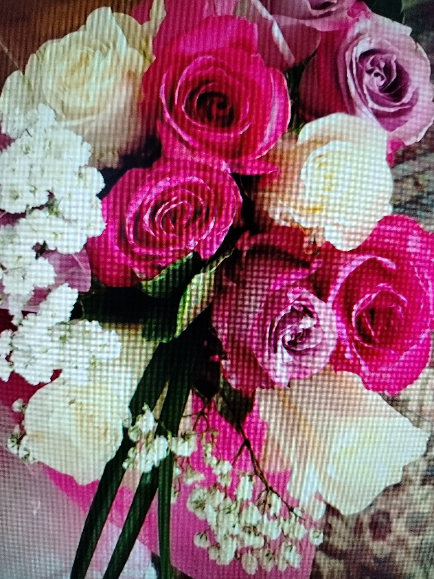 The image displays a close-up, overhead view of a floral arrangement. The bouquet is predominantly composed of roses in varying shades of pink, ranging from a deep fuchsia to a softer lilac. Interspersed with the roses are smaller, delicate white flowers, likely baby's breath, and some green foliage. The background appears to be a textured surface, possibly a fabric or rug, with a pattern in muted tones. There are no people or discernible actions taking place. The lighting is soft, highlighting the textures and colors of the flowers. The location is not specified within the image itself, and therefore cannot be identified as Yashkino, Russia or any other city.