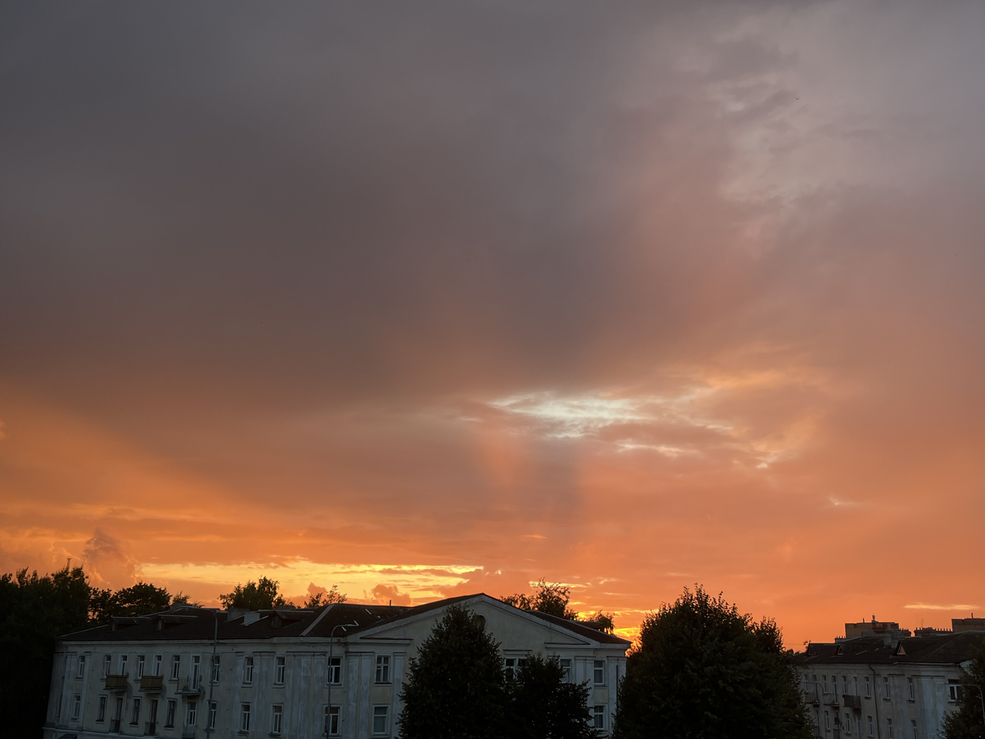 The image shows a dramatic sunset over a cityscape, likely in Kohtla-Järve, Estonia. The sky is a vibrant canvas of oranges, reds, and purples, with clouds creating interesting patterns. Some clouds are illuminated by the setting sun, while others are silhouetted against the brighter sky. In the foreground, several buildings are visible, characterized by their light-colored facades and dark roofs. Trees, also dark and lush, are interspersed among the buildings and along the horizon. The overall impression is one of a serene yet powerful natural event unfolding over an urban environment.