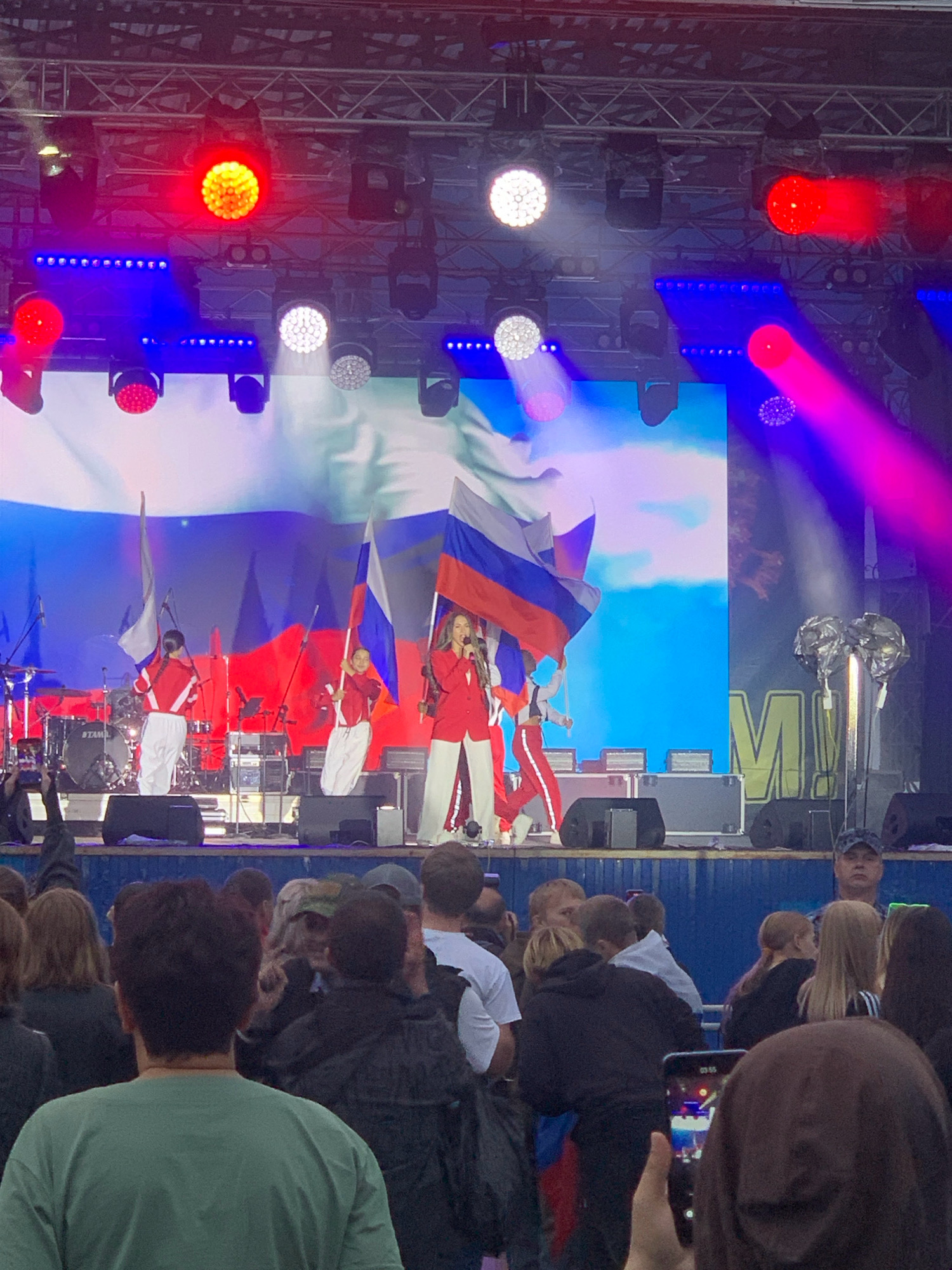 The image depicts an outdoor concert scene with a stage, performers, and an audience. The stage is lit with various colored lights, including red, blue, and white. In the background, a large screen displays a visual that appears to be a sky with clouds.
On stage, there are several individuals. A woman in a red jacket and white pants is singing into a microphone, positioned on the right side of the stage. She has long, blonde hair. Two other figures, likely performers or dancers, are also on stage. One is on the left, and another is in the center, both wearing matching red jackets and white pants, and holding Russian flags. Behind them, musical instruments, including drums, are visible.
The audience is in the foreground, with many people facing the stage. Some individuals are holding up phones, presumably to record the performance. The overall atmosphere appears festive, with the presence of flags suggesting a patriotic or national event.
The event appears to be taking place in the city of Prokopyevsk, Russia, given the presence of Russian flags and the context of the performance. The setting is outdoors, possibly a park or public square, with stage lighting and equipment set up for a performance. The weather seems to be overcast or late evening, as indicated by the artificial lighting.