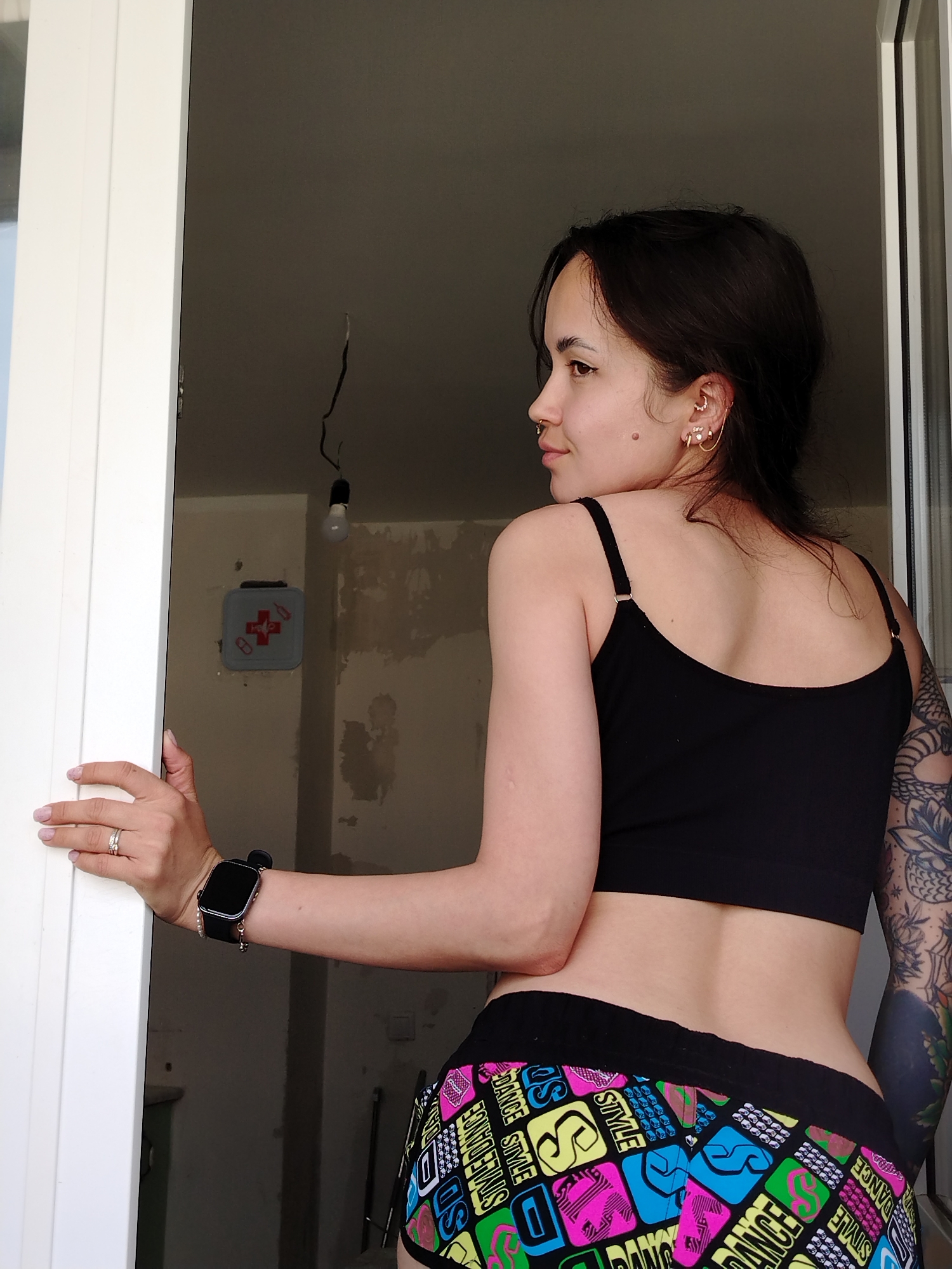 A young woman is captured from a rear-side perspective, standing in a white doorway or large window opening, looking over her left shoulder towards the left of the frame. Her dark hair is pulled back, revealing multiple intricate piercings on her right ear, including a chain cuff, and a nose ring. A small mole is visible below her right ear. She wears a black spaghetti strap crop top and colorful, patterned shorts with a black waistband. The shorts feature repeated words and symbols like "DANCE," "STYLE," and "SD" in neon pink, blue, green, and yellow squares. A large, dark tattoo covers her left upper arm and shoulder.
On her left hand, she wears two silver rings on her ring finger, and a black smartwatch with a small pearl charm is on her left wrist. Her fingernails are neatly manicured with light polish.
The background reveals an unfinished room, likely under renovation. The walls are bare, showing patches of light gray plaster or concrete. Exposed wiring hangs from the ceiling, with a single bare light bulb. A small gray first aid kit box with a red cross symbol and the word "HELP" is mounted on the wall to the left of the light. Natural light appears to be entering from outside the opening, suggesting it is daytime. The scene conveys a moment of pause or observation within a construction or renovation environment.