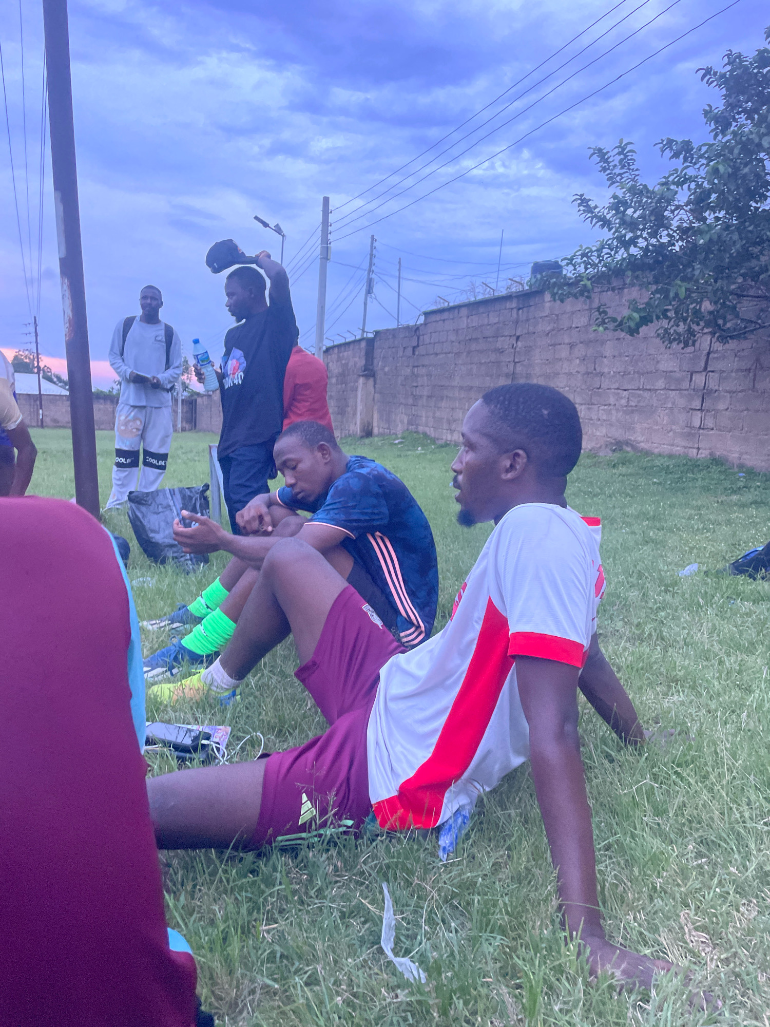 The image depicts a group of young men sitting on a grassy field, appearing to be taking a break. The setting is likely an outdoor sports facility or a park in Jos, Nigeria, given the overcast sky and the presence of a brick wall in the background, characteristic of such environments.
In the foreground, two individuals are prominently visible. One, wearing a white t-shirt with red accents on the sleeves and collar, is seated with his legs extended. His skin tone is dark, and he has a short haircut with a goatee. To his left, another individual in a dark blue t-shirt with red stripes on the sleeves is also seated, leaning slightly forward. He has bright green socks pulled up to his knees and is wearing athletic shoes. His hands are resting on his knees.
Behind them, another man in a black t-shirt is standing and holding a black baseball cap in his raised right hand. He is holding a clear plastic bottle of water in his left hand. To his left, another man in a light-colored long-sleeved shirt and grey athletic pants with graphic prints is also standing, looking downwards. Further in the background, a man in a red shirt is partially visible.
Other objects in the scene include a black trash bag on the ground near the standing men, and a phone plugged into a power bank, also on the grass near the seated individuals. The overall atmosphere suggests a pause during physical activity, perhaps a sports practice or game. The lighting indicates it is likely late afternoon or early evening, with the sky a mix of grey and pale blue, suggesting an impending dusk or a cloudy day.