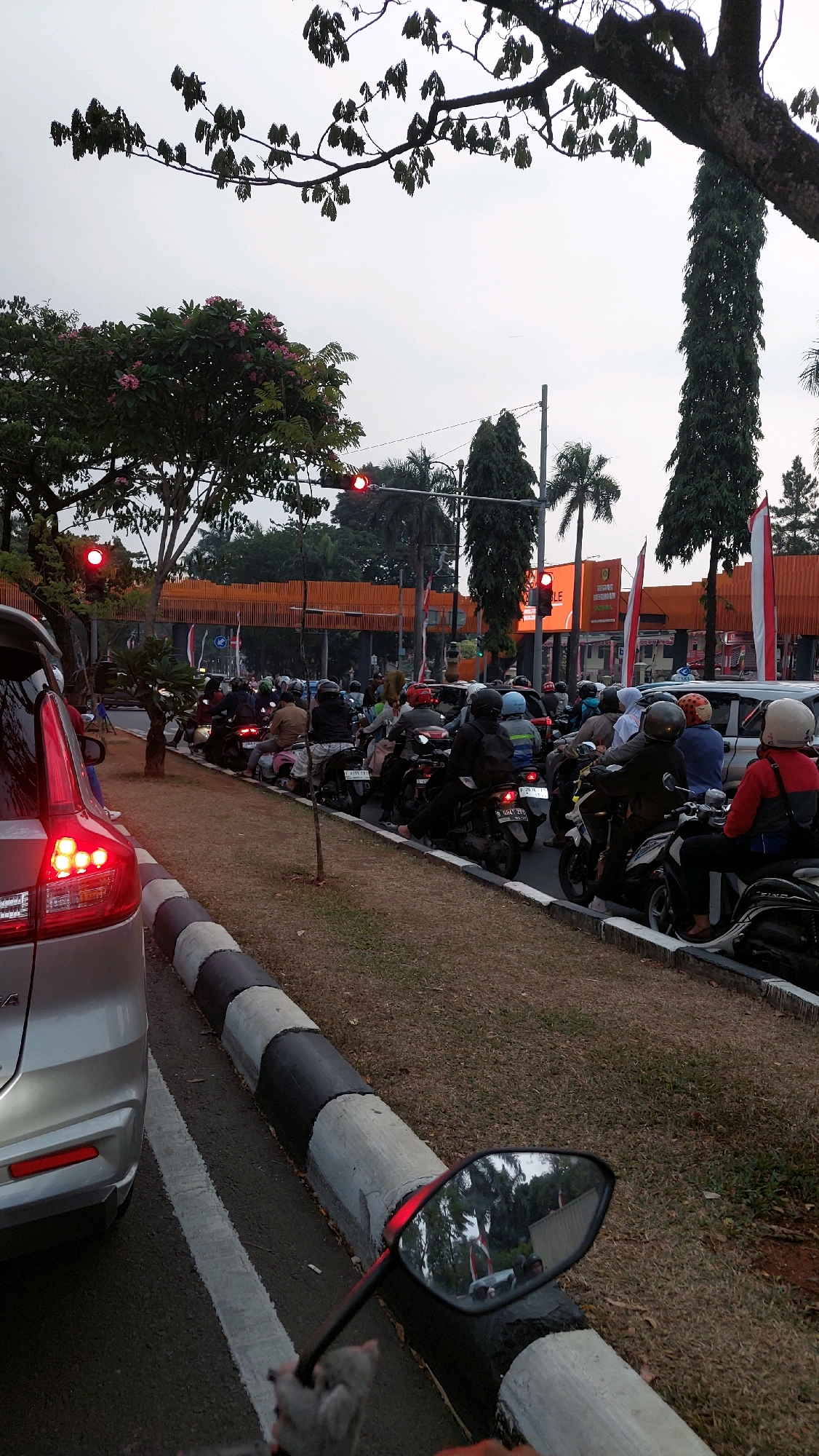 This is a photograph taken from a motorcycle in Cibinong, Indonesia, capturing a traffic jam scene. The foreground features a silver car's rear, its taillights illuminated red. To the right, the motorcycle's side mirror reflects the road and other vehicles. The midground shows a long line of motorcycles, many with riders wearing helmets, queuing on the road. Behind the motorcycles, traffic lights are visible, one displaying red. The background reveals a pedestrian bridge with orange accents, buildings, palm trees, and other foliage, indicating an urban environment. The sky is overcast.