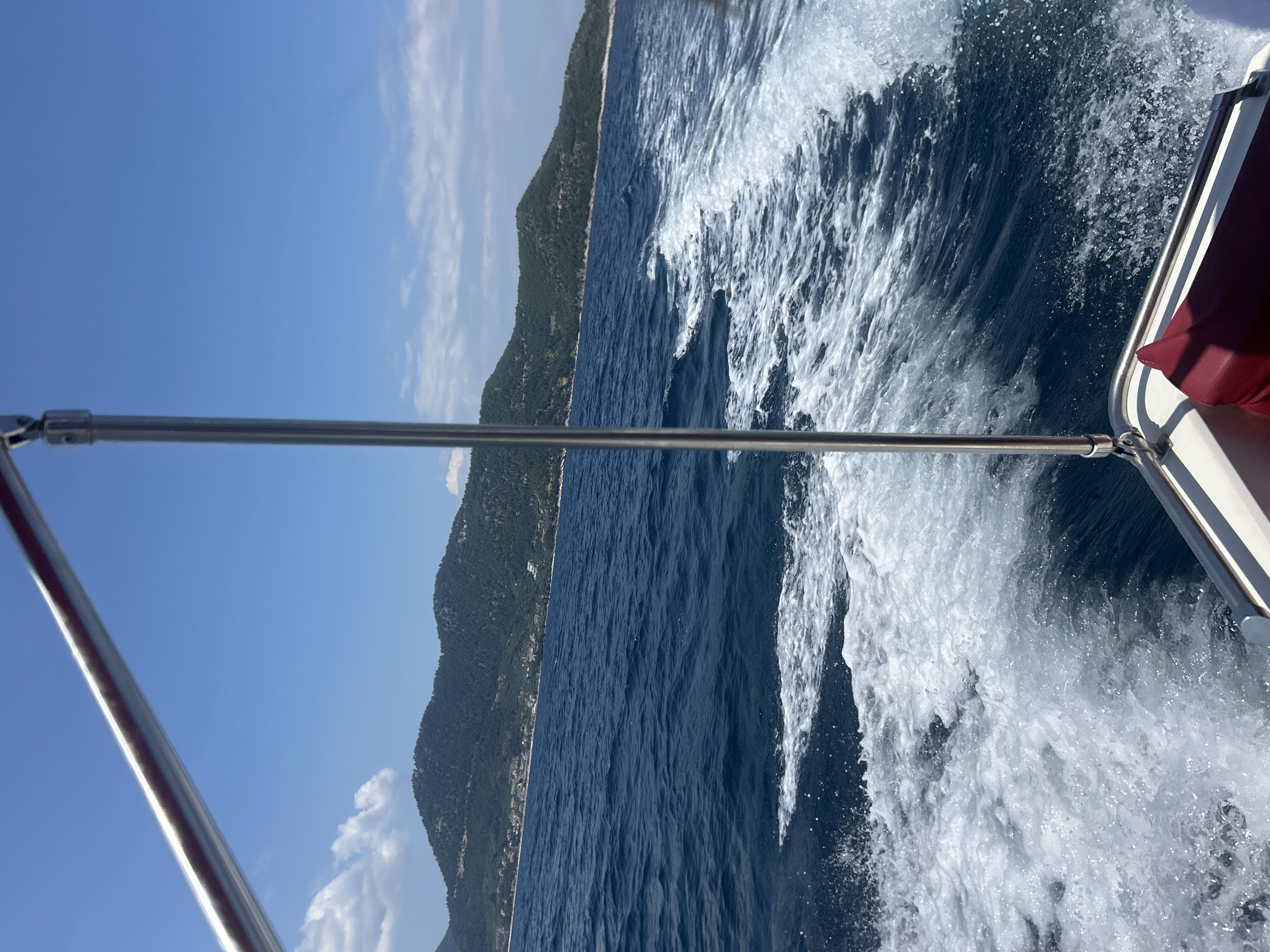 The image depicts a view from the stern of a boat moving through deep blue water, creating a white wake. The scene is characterized by a clear blue sky with scattered white clouds. In the background, a lush green, tree-covered island with a small village nestled along its coast is visible. The boat appears to have a stainless steel railing and a red cushion is partially visible in the lower right corner. The vantage point is from a moving vessel, suggesting a journey or leisure activity on the water.