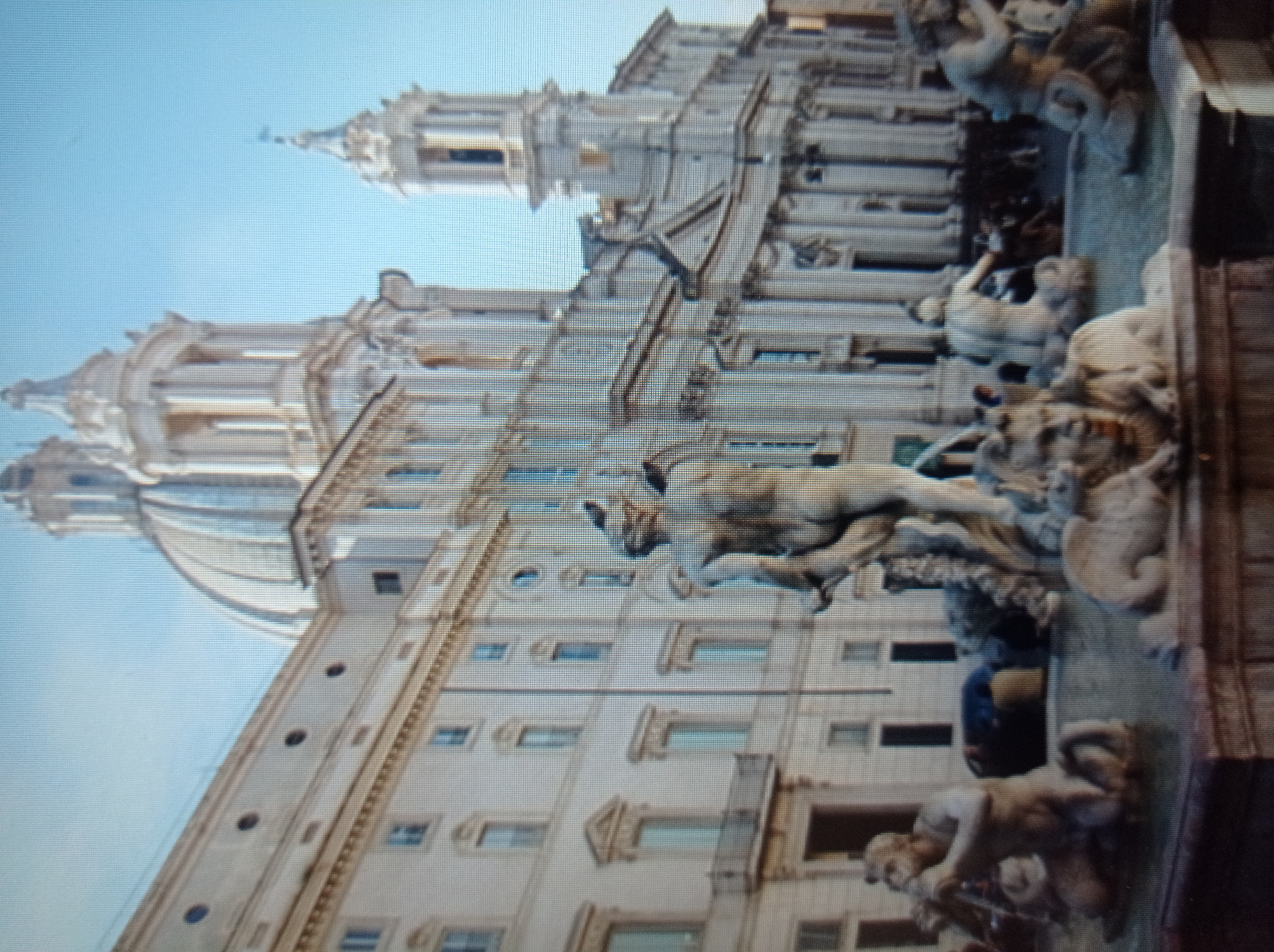 The image shows a wide view of a large, ornate fountain in a city square. The fountain features several sculpted human figures and possibly mythical creatures. In the background, a grand, pale building with a large dome and multiple towers dominates the skyline. The architecture suggests a European city. The sky is a clear, light blue. There are a few indistinct figures of people visible in the background, near the fountain and the building. The image is somewhat blurry, and the details are not sharp.
The image does not depict any recognizable landmarks or characteristics of Mukachevo, Ukraine. Instead, the architectural style of the building and the design of the fountain are strongly indicative of a location in Italy, possibly Rome.