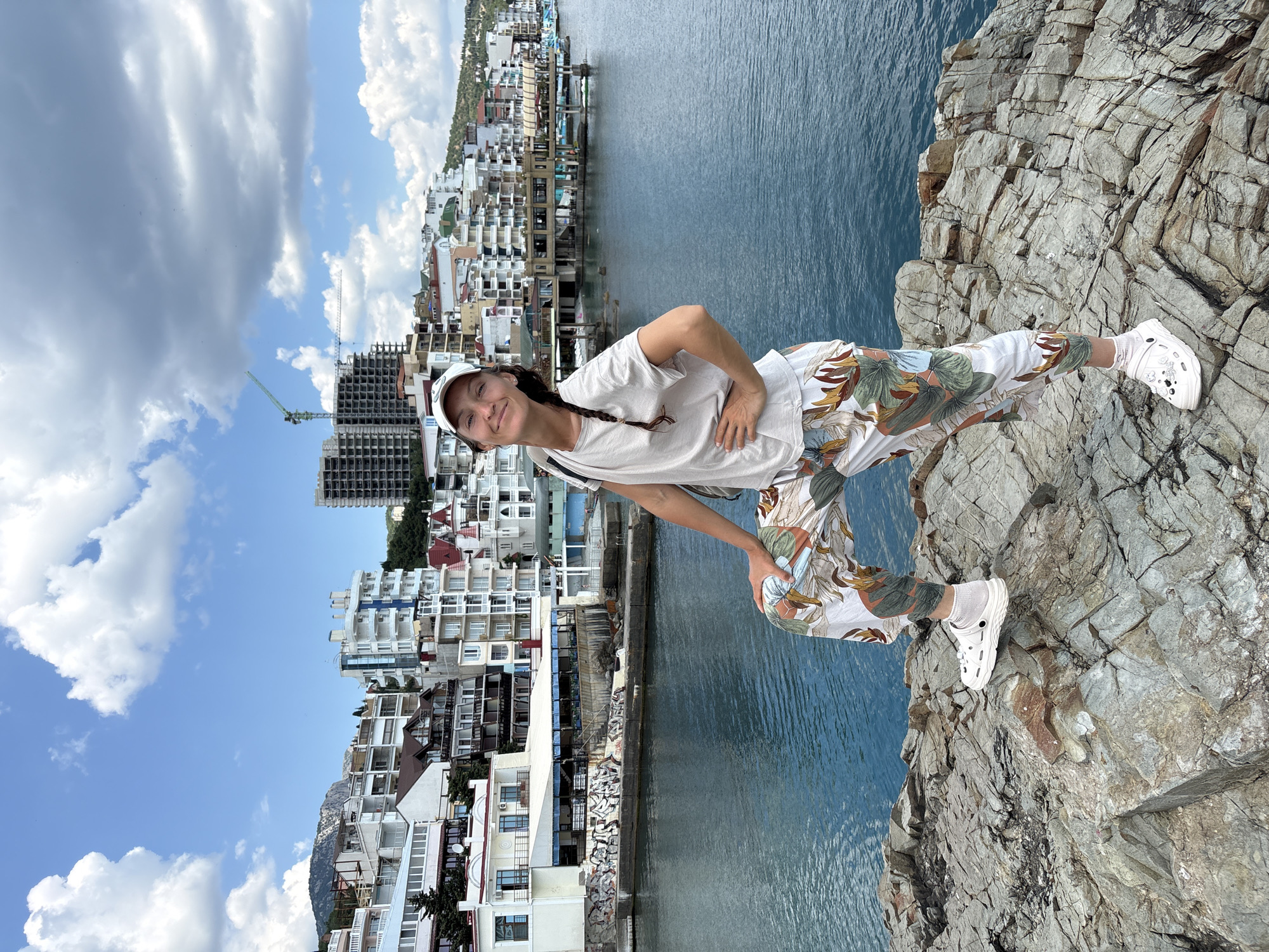 A full-body, medium shot captured outdoors on a sunny day shows a woman standing on rocks with a body of water and a city in the background. The woman, positioned slightly right of center, faces forward and slightly to the left, her head tilted and a smile on her face. She wears a white baseball cap, a light-colored t-shirt, and white pants with a tropical leaf print. Her right hand rests on her hip, and her left leg is bent and raised, with her foot resting on a rock. She is wearing white clogs with white socks. The background features a body of blue water. Beyond the water, a coastal city is visible, characterized by multi-story buildings with white facades and balconies, and some under construction. The sky above is blue with scattered white clouds. The foreground consists of textured gray rocks.