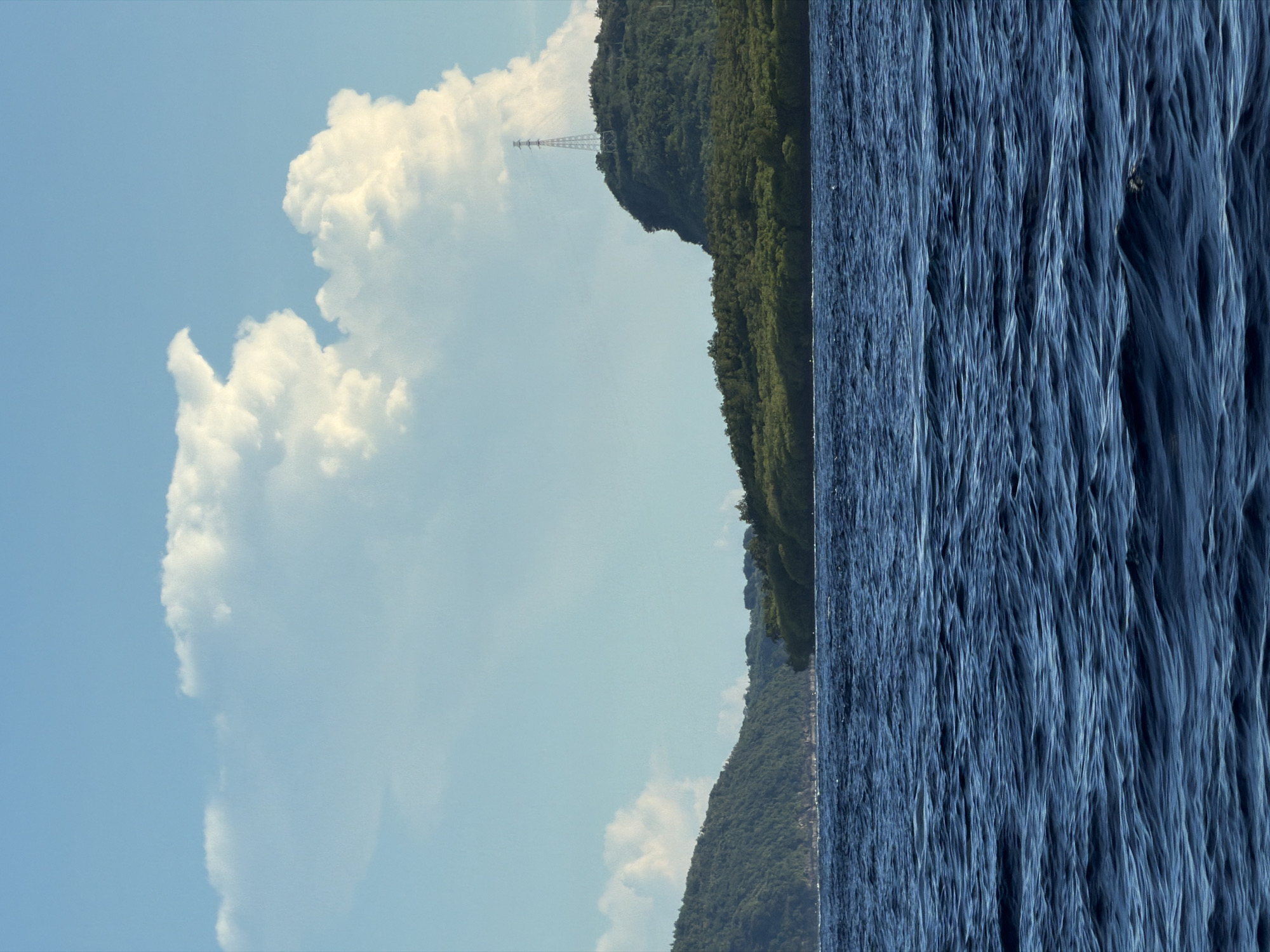 The image displays a serene natural landscape featuring a body of water, a forested mountain range, and a cloudy sky, presented as if rotated 90 degrees clockwise to a horizontal orientation. The upper part of the frame is dominated by a clear, light blue sky, adorned with large, fluffy white cumulus clouds. A significant cloud mass occupies the upper left portion, showcasing its distinct textures.
Across the mid-ground, a densely forested mountain range forms a natural horizon line, separating the sky from the water. The mountains are covered in lush, dark green foliage. On one of the prominent peaks, a tall, slender metal lattice tower, likely a telecommunications or transmission structure, stands silhouetted against the bright sky.
The lower section of the image is filled by a wide expanse of deep blue water, which appears to be either a lake or a sea. The water's surface is characterized by numerous small, parallel ripples and waves, creating a dynamic texture of light and shadow across its entirety.
The scene depicts a calm, bright daylight setting, suggesting late morning or early afternoon. The weather is clear with visible clouds. No people or discernible text are present in the image.