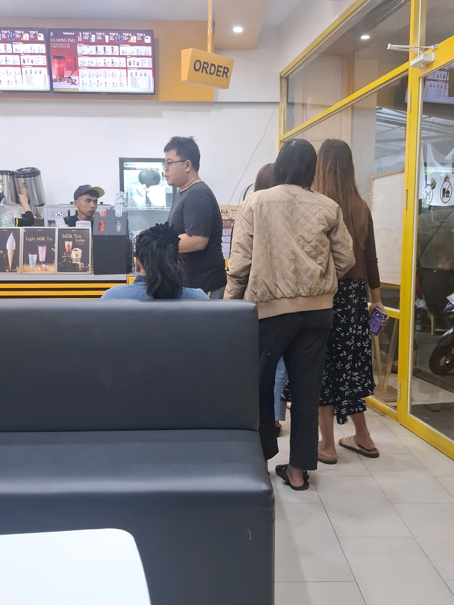 The image shows the interior of a fast-food establishment, likely a bubble tea or beverage shop, in Cianjur, Indonesia. A yellow sign with the word "ORDER" is visible, indicating the ordering area. Several people are present. Two employees are behind the counter, one in a black t-shirt and a baseball cap, and another partially visible on the left. A customer, wearing a denim jacket and with an elaborate updo, is seated on a dark gray bench, facing away from the camera, looking towards the counter. In front of her, three customers are standing in line. One customer in a light brown quilted jacket and black pants is facing the counter. Next to him, two women are standing; one with long brown hair wearing a dark top and a patterned skirt, holding a purple phone, and another person whose face is obscured. The environment appears clean and brightly lit, with tiled flooring. Menus and promotional displays are visible on the wall behind the counter. The overall scene depicts a typical customer interaction at a food service location.