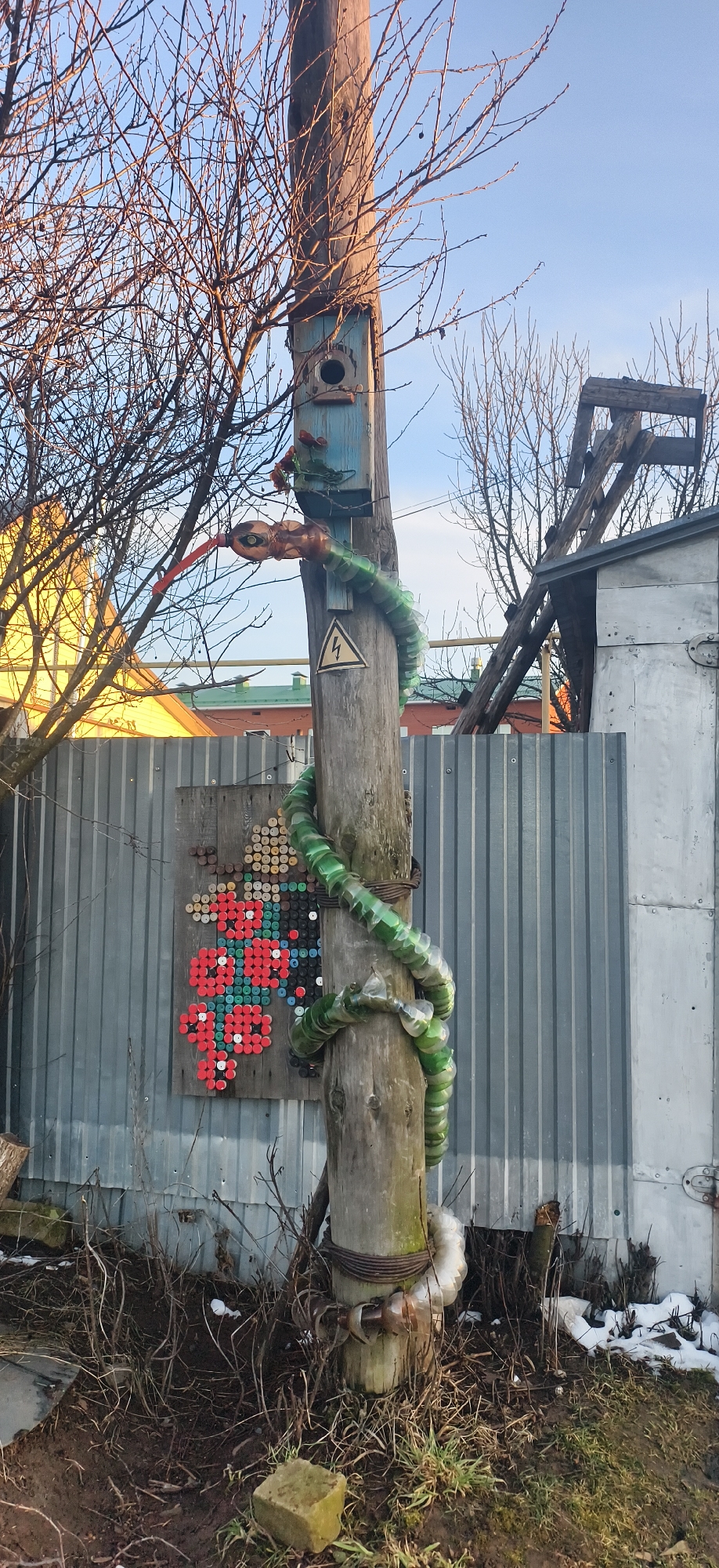 The scene is an outdoor setting, likely in a residential or semi-rural area of Фоки, Russia. A weathered wooden utility pole stands as the focal point. A handcrafted snake, constructed from green plastic bottles, is coiled around the pole. The snake's head appears to be made from a clear plastic bottle, with a red plastic utensil serving as its tongue. Above the snake, a small, blue-painted birdhouse is attached to the pole. A yellow "high voltage" warning sign is affixed to the pole.
Behind the pole, a corrugated metal fence spans the background. Affixed to the fence, to the left of the pole, is a piece of artwork constructed from colorful bottle caps, depicting a floral design. To the right of the pole and fence, a dilapidated wooden structure is visible. Bare tree branches are visible in the background.
The ground at the base of the pole is covered with sparse vegetation, dead leaves, and small patches of snow. A weathered stone is visible at the base of the pole.