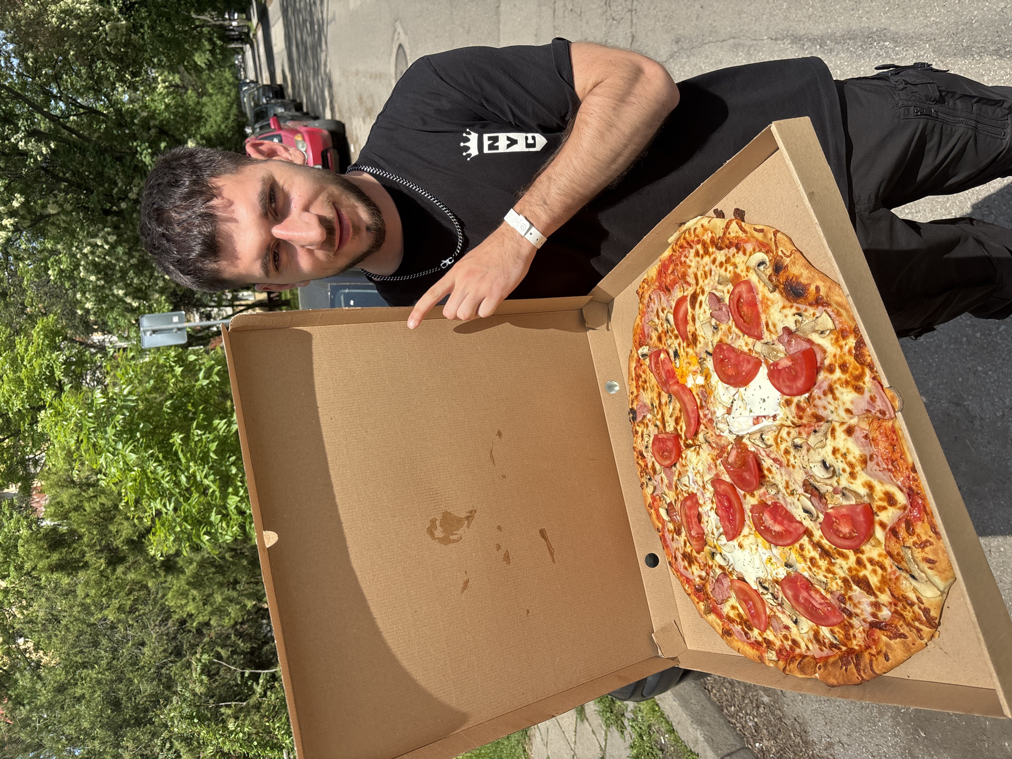 A man stands outdoors, pointing at a pizza in a pizza box. The pizza is topped with tomatoes, cheese, and mushrooms. He is wearing a black t-shirt, black cargo pants, and a silver chain necklace. He has a white wristband on his left wrist. The background includes trees, a street, and parked cars. The location is in a city, the specific name of which is unknown.
