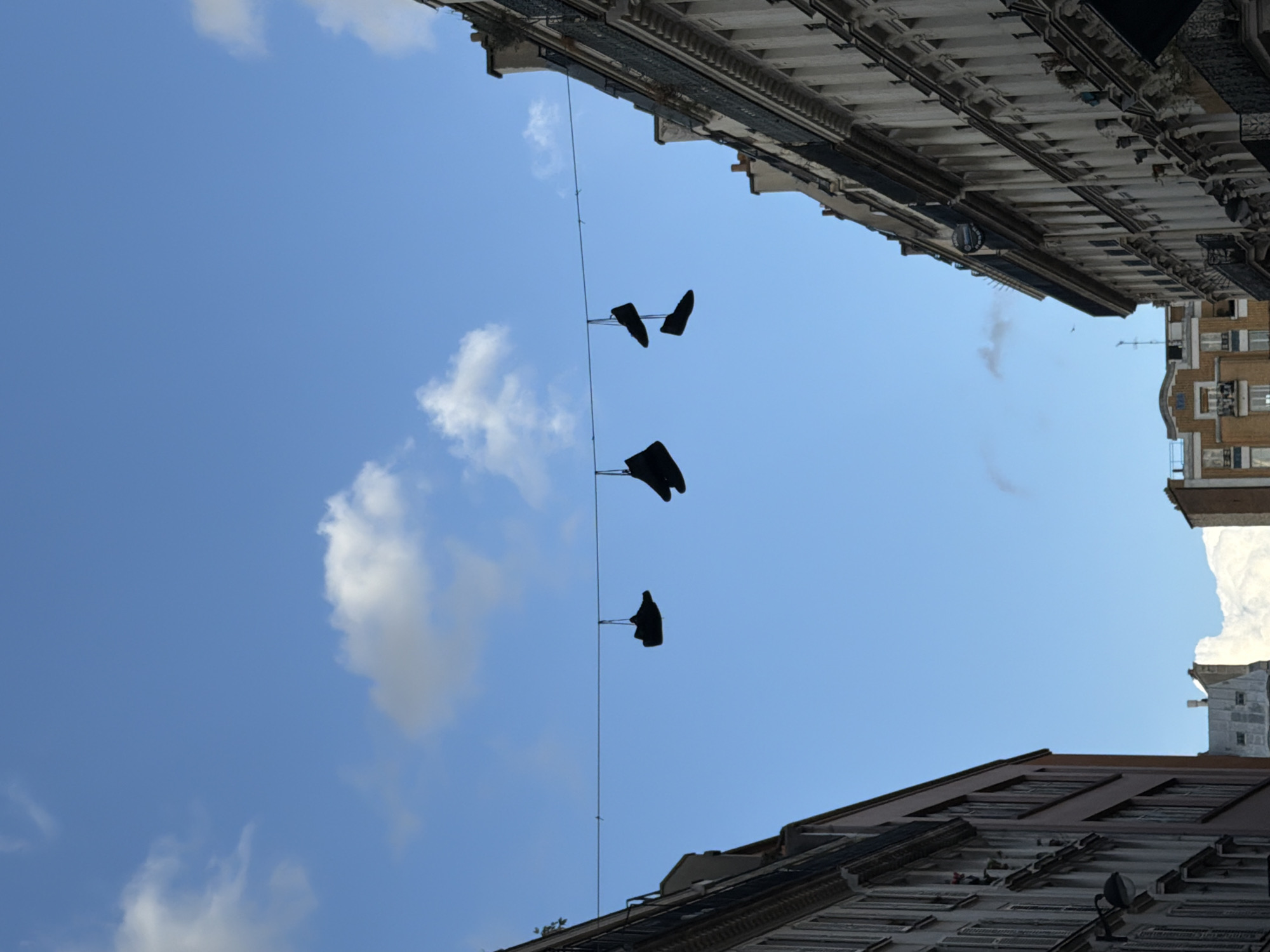 The image captures a view looking upwards in Paris, France. At the top and bottom of the frame, the intricate architectural details of Parisian buildings are visible. A wire stretches across the sky between the buildings, from which three pairs of shoes are suspended. The sky is blue with some white clouds.