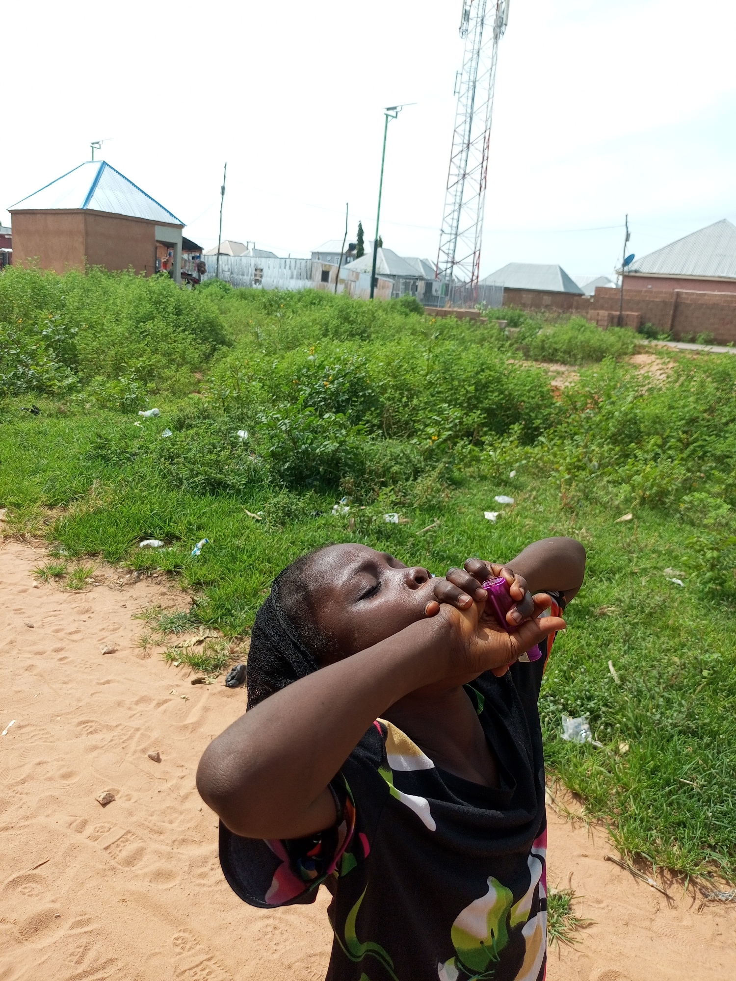A young Black girl, wearing a black headscarf and a black t-shirt with a floral pattern, is outdoors in a sandy area with green vegetation behind her. The girl has her eyes closed and is holding a small purple object to her lips as if blowing into it. In the background, there are buildings, a tall cell tower, and scattered white plastic bottles and wrappers in the green grass. The scene appears to be in a rural or semi-urban area, possibly a village or outskirts of a town. The overall impression is that of an everyday moment in a developing area. The image is well-lit and appears to have been taken during daylight hours.