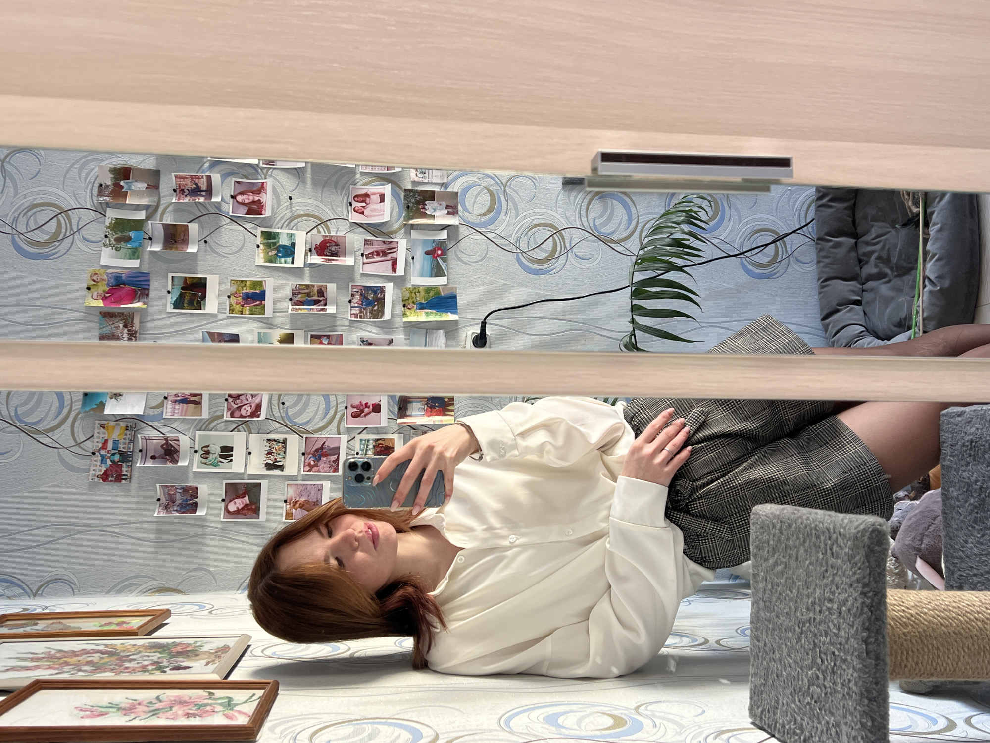 The image depicts a young woman taking a mirror selfie within what appears to be a domestic interior in Chaikovsky, Russia. The woman, with shoulder-length reddish-brown hair, is wearing a loose-fitting white collared shirt and plaid shorts. She holds a light blue or green smartphone with her right hand, positioned to capture her reflection in a full-length mirror, which is partially obscured by a light-colored wooden door frame on the right. Her left arm rests against her side, and a ring is visible on her left hand.
She is leaning against a wall covered in light blue and grey wallpaper featuring abstract circular patterns. To her left, two framed pictures depicting floral art are partially visible on the wall. Below her, a grey, carpeted cat tree with rope-wrapped posts is present. In the background of her reflection, numerous small photographs are affixed to the wallpaper, seemingly creating a collage held by thumbtacks and possibly interconnected by a string. A black electrical cord plugs into an outlet on this wall and extends downwards, partially obscuring a leafy green houseplant. On the floor in the reflected background, a grey pet bed is visible. The lighting suggests an indoor setting, likely a room within a residence.