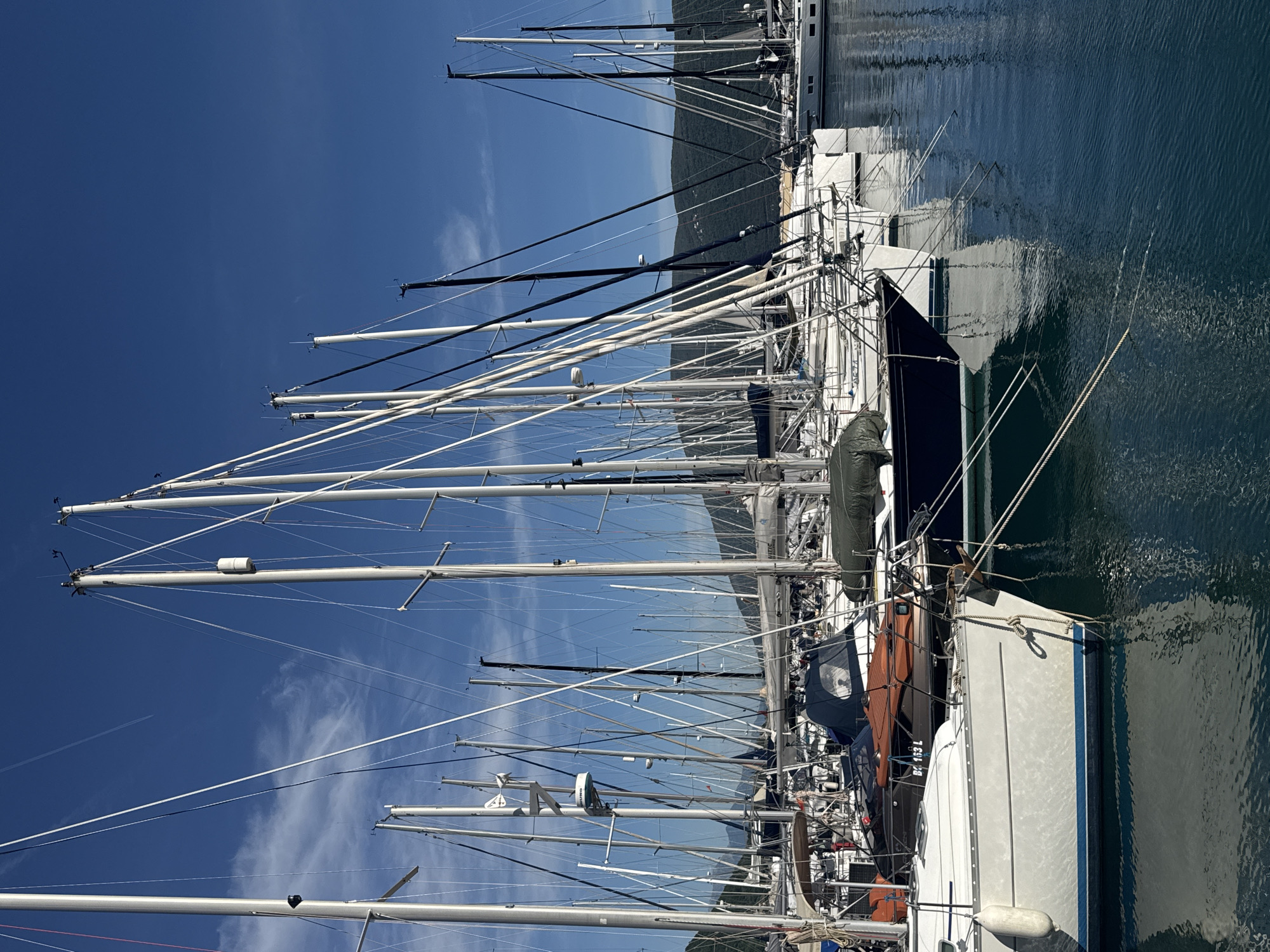 The image depicts a marina in Meljine, Montenegro, filled with yachts. The composition focuses on the masts of numerous sailboats docked side-by-side in the harbor. The masts are predominantly white and reach towards a clear, blue sky with scattered cirrus clouds. The masts create a dense network of vertical lines, contrasting with the blue sky and water. The hulls of the boats are visible in the foreground and middle ground, with varying colors and details. The water reflects the sky and the hulls of the boats, creating a mirror-like effect. In the background, there is a hilly landscape covered with vegetation.
