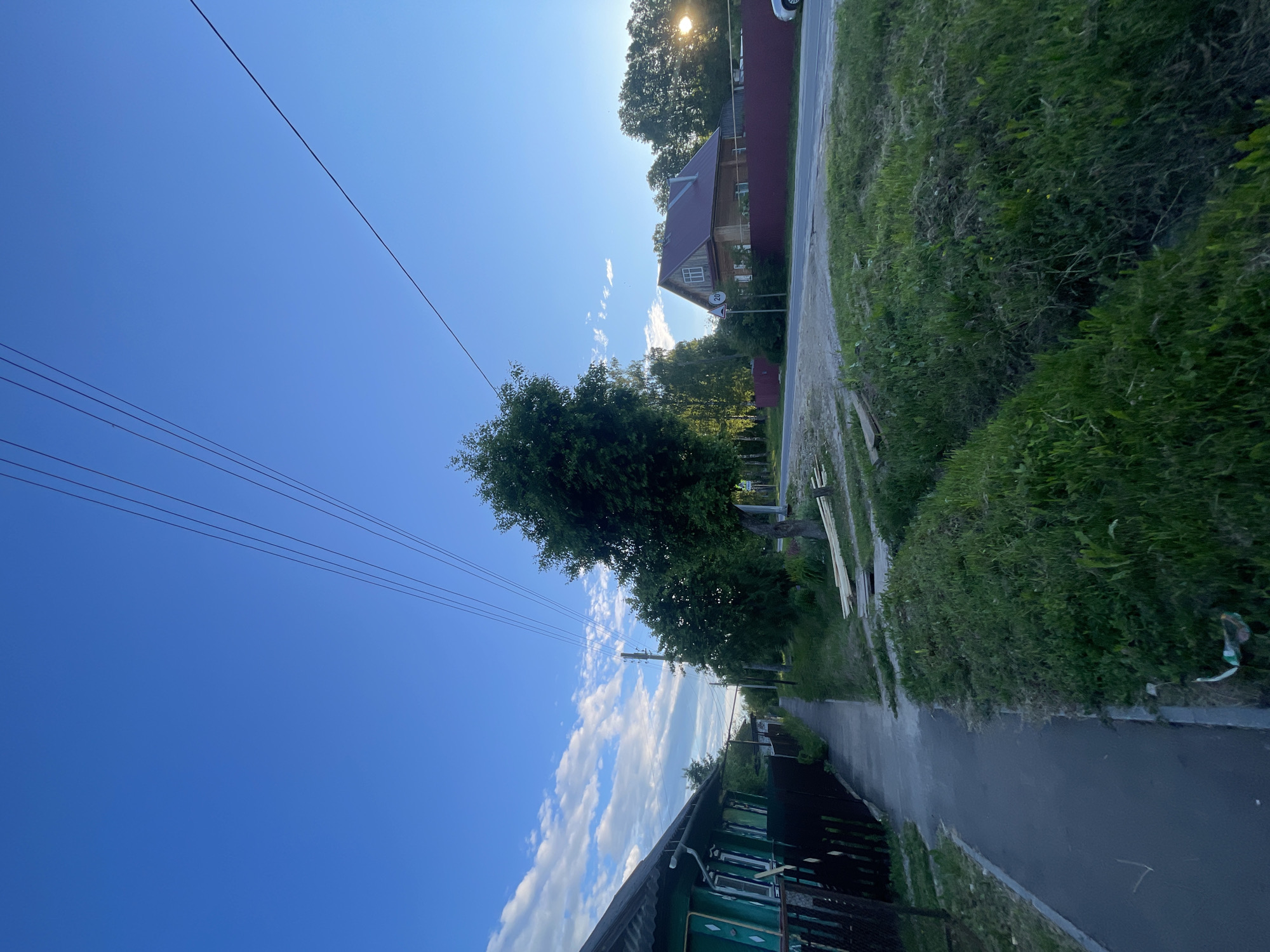 The image depicts a street scene in the settlement of Krasnye Baki, Russia. On the left side of the frame, a green wooden house with a dark roof and fence runs along a paved sidewalk. The sidewalk leads into the distance, bordered by lush green grass on the right. Further down the street, several trees line the path, and a wooden utility pole supports overhead wires. On the right side of the street, across from the sidewalk, is a residential building with a red roof, surrounded by trees and a dark red fence. A speed limit sign with the number "20" is visible near the house. The sky is a clear, vibrant blue with a few scattered white clouds. The sun is setting or rising, casting a warm glow and a lens flare in the upper right portion of the image. The overall impression is a quiet, rural or semi-rural setting in the late afternoon or early morning. There are no people visible in the image.