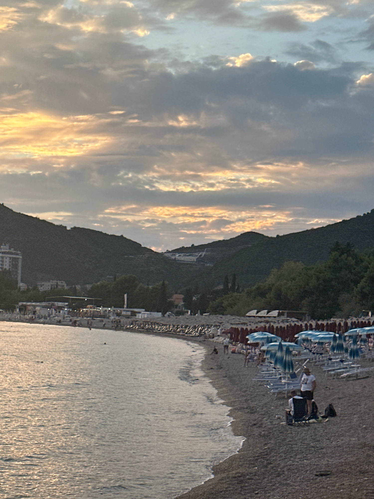 The image depicts a beach scene in Budva, Montenegro, during what appears to be late afternoon or early evening, judging by the soft lighting and cloud formations. The sky is a mix of gray and yellow, with sunlight breaking through the clouds, creating a warm glow.
The beach itself is covered in dark pebbles. In the foreground, a calm sea laps gently at the shore. The middle ground shows a populated beach with numerous rows of lounge chairs and umbrellas, predominantly in blue and white striped patterns, set up for beachgoers. Several people are visible on the beach, some sitting in chairs, others standing, and a few are walking. Two individuals are prominently featured in the lower right, one standing in a white t-shirt and dark shorts, and another seated in a foldable chair.
The background is dominated by lush green, tree-covered hills that rise steeply behind the beach. A tall, modern building, likely a hotel, is visible on the left side of the image, nestled into the hillside. Further back, on the slopes of the hills, structures and roads are faintly discernible, suggesting the presence of a town or resort area. The overall impression is that of a lively, scenic beach location on the Adriatic coast.