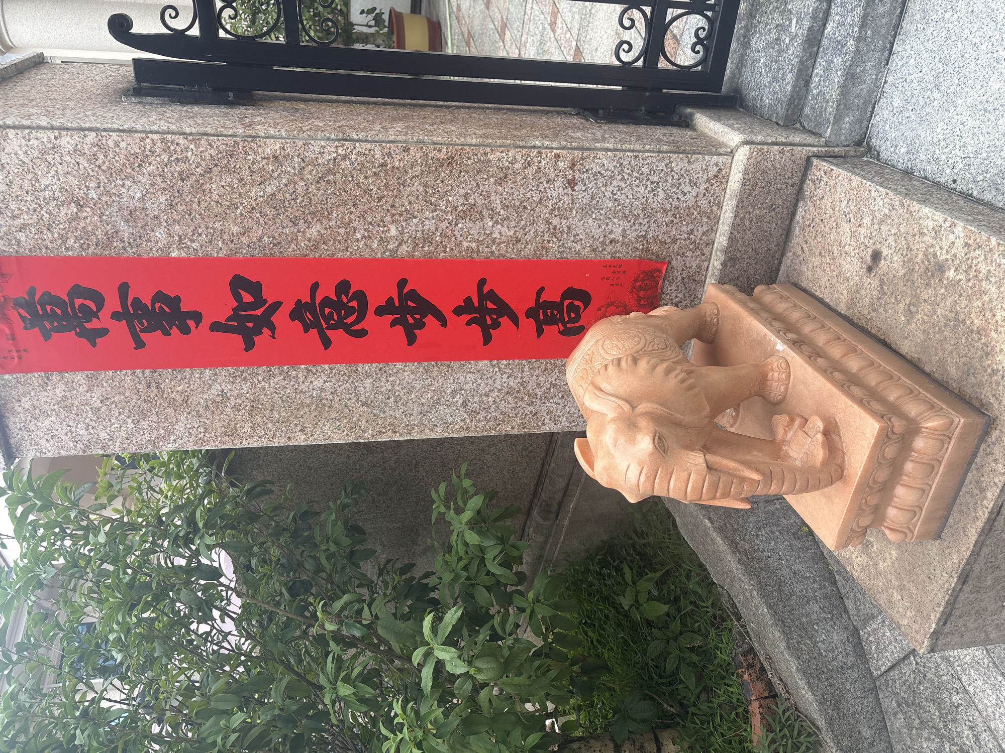 In Nanhai District, China, a stone elephant statue stands on a decorative pedestal in front of a granite-clad pillar. A red banner with black Chinese calligraphy hangs vertically on the pillar. To the right of the pillar, a black wrought-iron gate is visible. Lush green foliage from a nearby tree and ground cover surrounds the base of the pillar and extends to the left of the frame. The ground is paved with stone tiles.