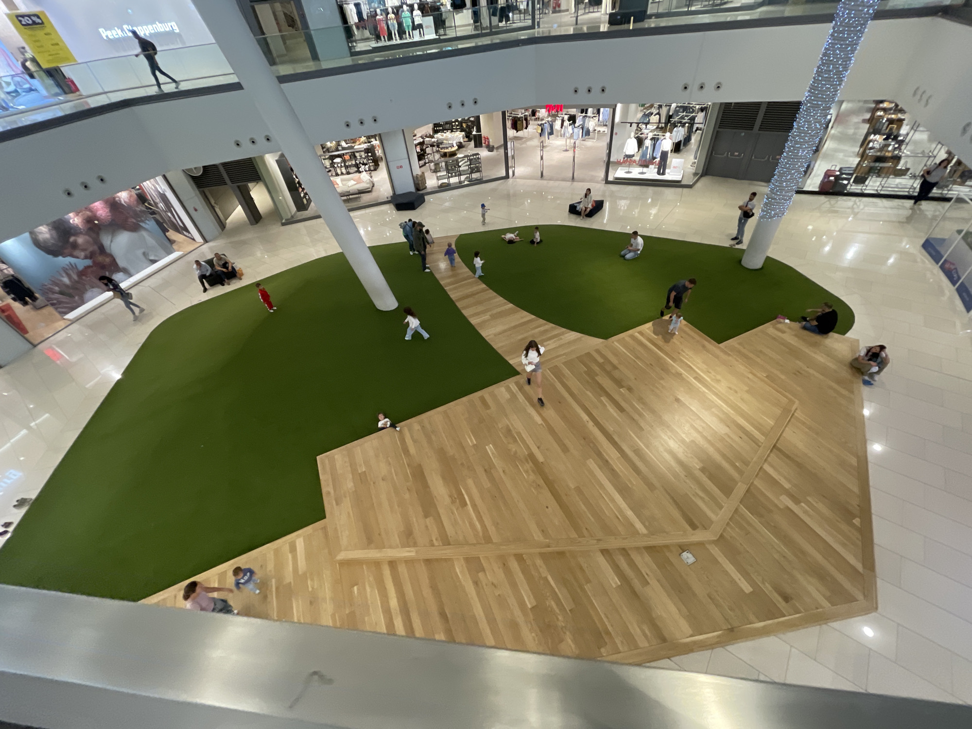 This is an aerial view of the interior of a multi-level shopping mall. In the foreground, a section of a wooden multi-tiered structure is visible, with people sitting on it. This structure is surrounded by a vibrant green, artificial grass area. Beyond this green space, several people, including children, are seen walking or standing. In the background, the mall's upper levels are visible, with various retail stores displaying clothing. The mall has polished floors and large white support columns. There is a decorative, illuminated pillar in the background. The scene appears to be a public, indoor recreational space within a commercial environment. This location is likely the mall "The Mall" in Sofia, Bulgaria, which is known for its architectural design featuring a green, tiered central area.