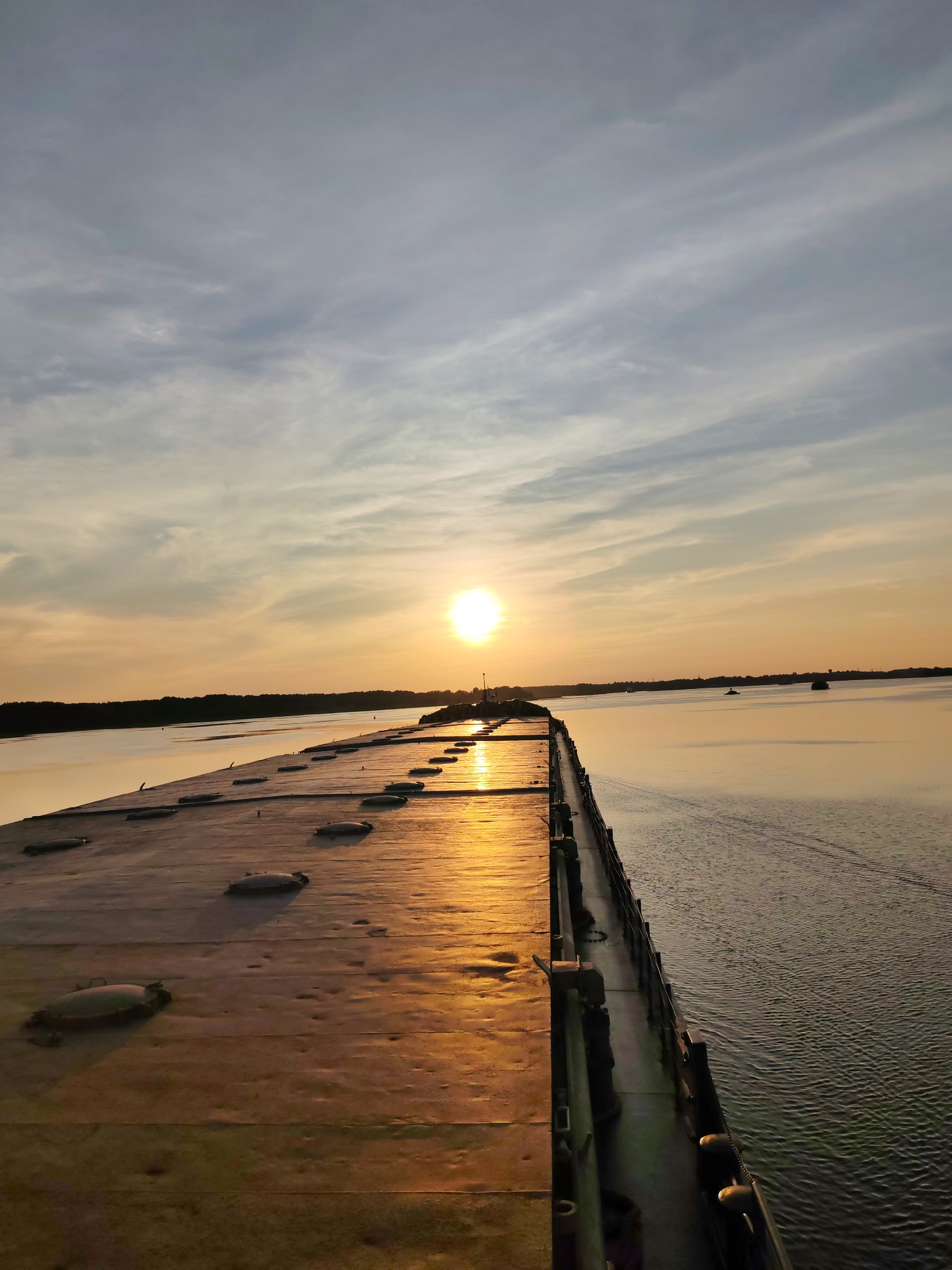 An outdoor, eye-level, medium shot captures a perspective view of a long, flat, metallic structure extending into a calm body of water. The sun, partially obscured by clouds, casts a warm, golden glow across the scene, reflecting brightly on the metallic surface and the water. The sky is filled with scattered, wispy clouds, and the horizon is marked by a dark, treeline. The metallic structure appears to be a pontoon or a floating platform, with numerous circular or oval hatches dotting its surface. The water is calm, with subtle ripples indicating slight movement. In the distance, on the water, a few small objects, possibly boats or buoys, are visible. The overall scene suggests an industrial or maritime environment, possibly a harbor or a section of a river or lake. The specific location is within the city of Shchetinino, Russia.